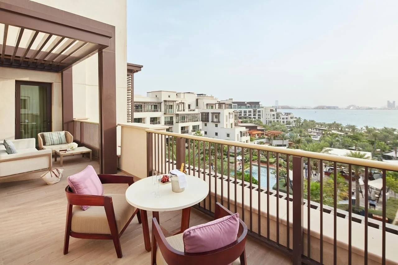 Balcony view of a resort with outdoor seating, a small round table with glasses and grapes, overlooking white modern buildings, a pool, palm trees, and ocean in the distance.