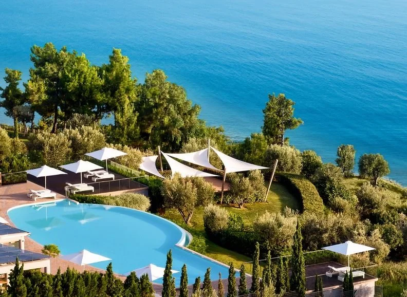 Pool area with white umbrellas, lounge chairs, and cabanas on a hillside overlooking the ocean, surrounded by greenery and trees.