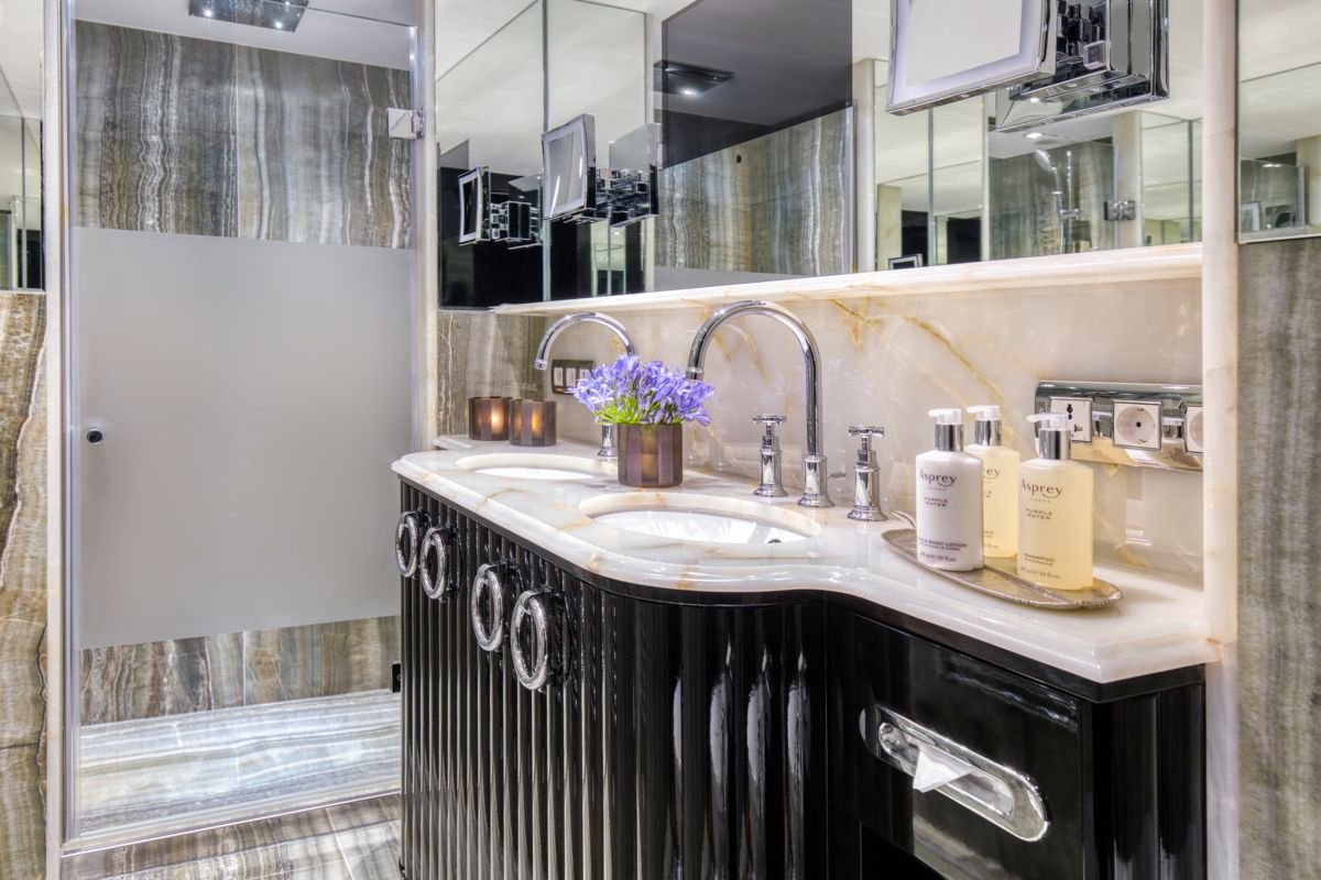 A luxurious bathroom vanity area with a marble countertop, three sinks, and a large mirror. A small flower arrangement and toiletries are on the counter, with mirrored cabinets and a glass shower door to the left.