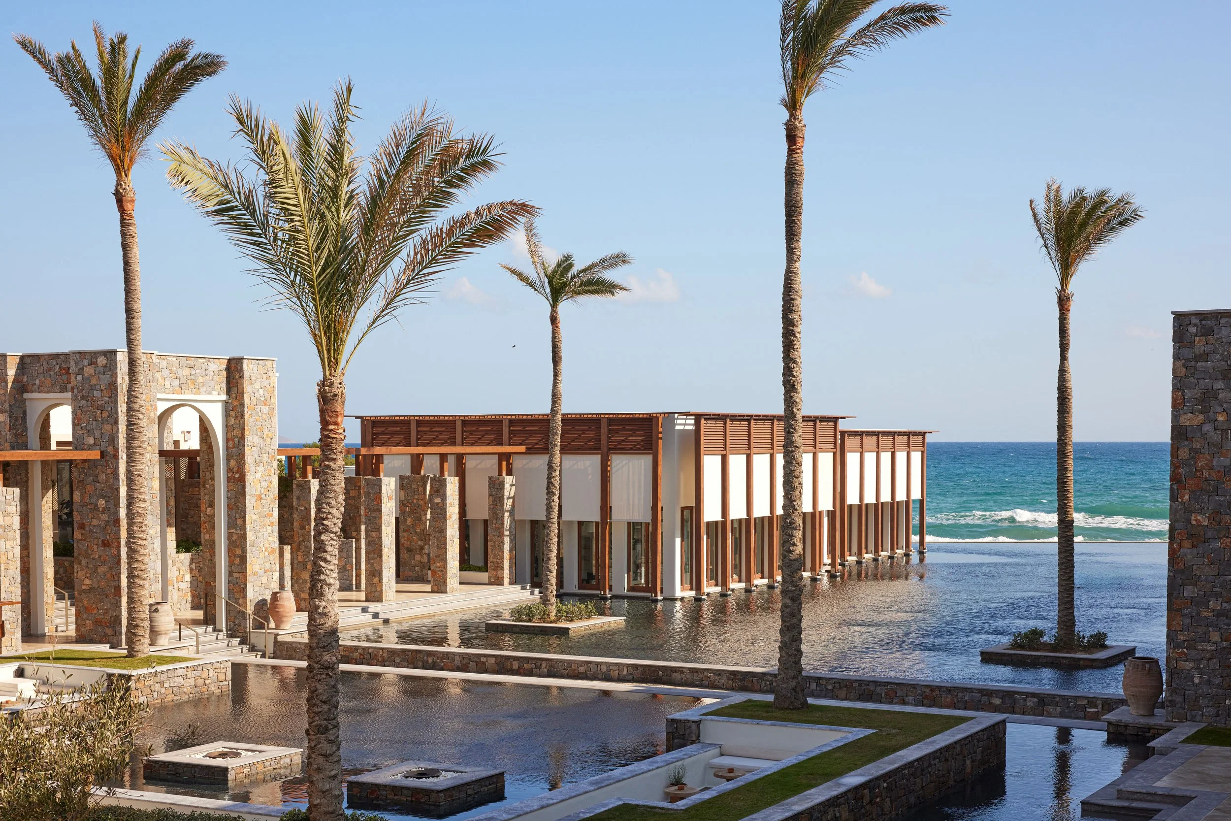 Modern beachfront building with stone and wood architecture, tall palm trees, water features, and ocean in the background under a blue sky.