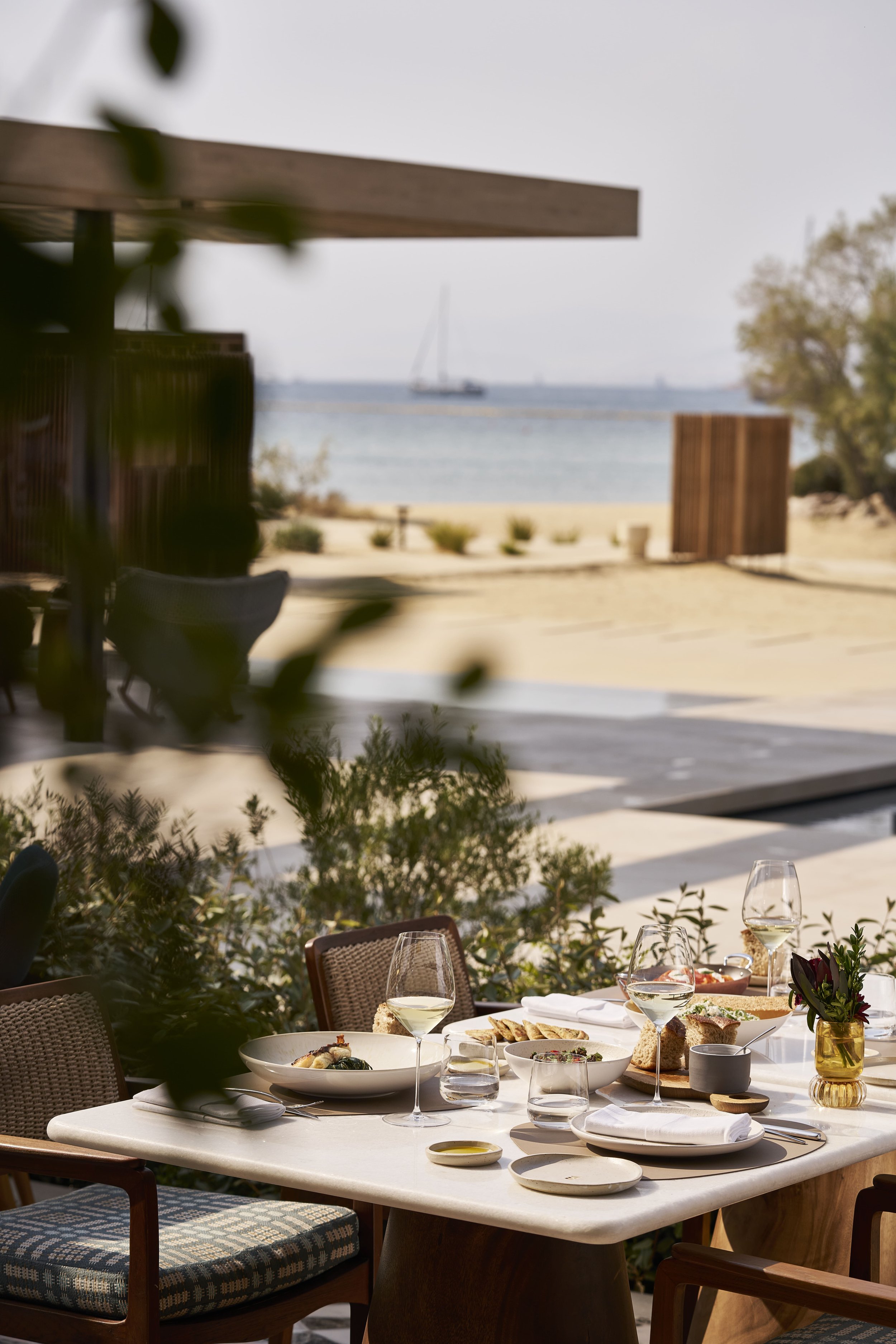 An outdoor dining scene with a table set with plates of food and glasses of white wine, overlooking a beach with sailboats in the water.