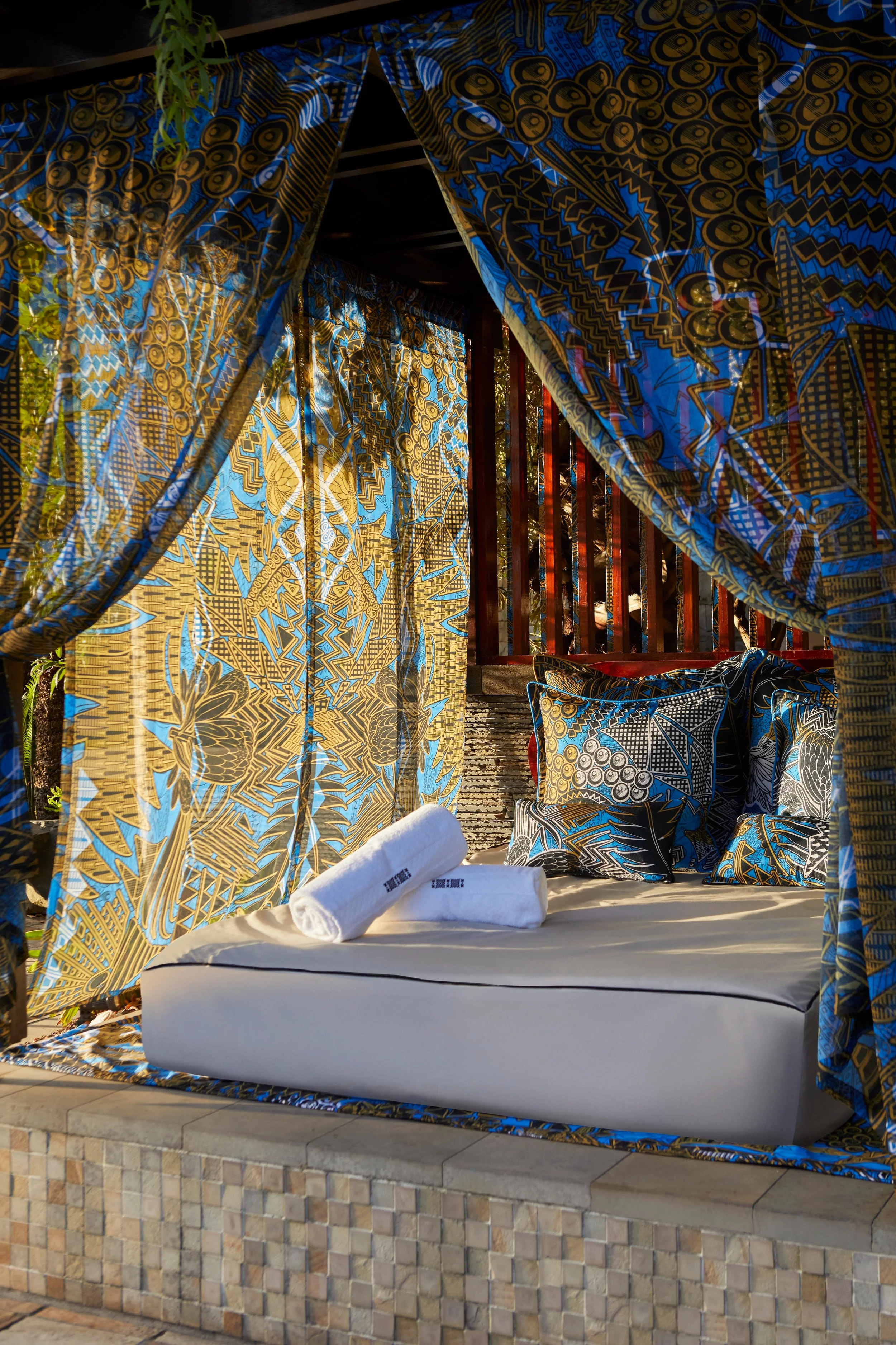 An outdoor cabana with colorful patterned curtains and cushions. A bed with white linens and rolled towels, set on a tiled platform.