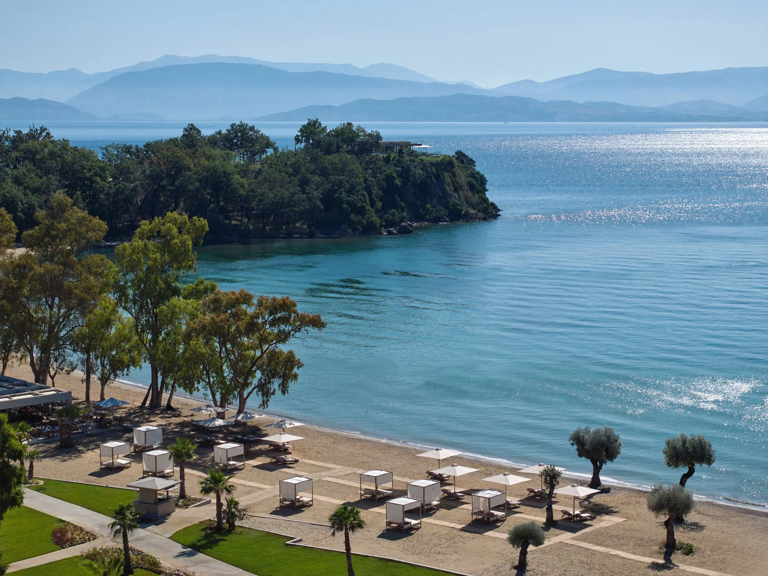 A scenic view of a beach with white umbrellas and shaded cabanas along the shore, lush trees, and a calm ocean with mountain ranges in the background under a clear blue sky.