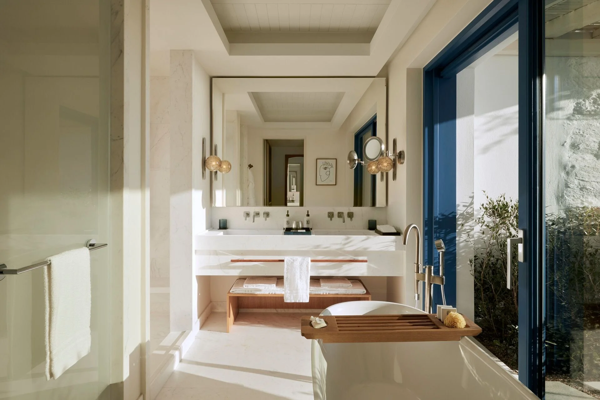 Modern bathroom with a large mirror above a white marble sink, decorative wall sconces, and blue-framed glass doors leading to an outdoor garden area.
