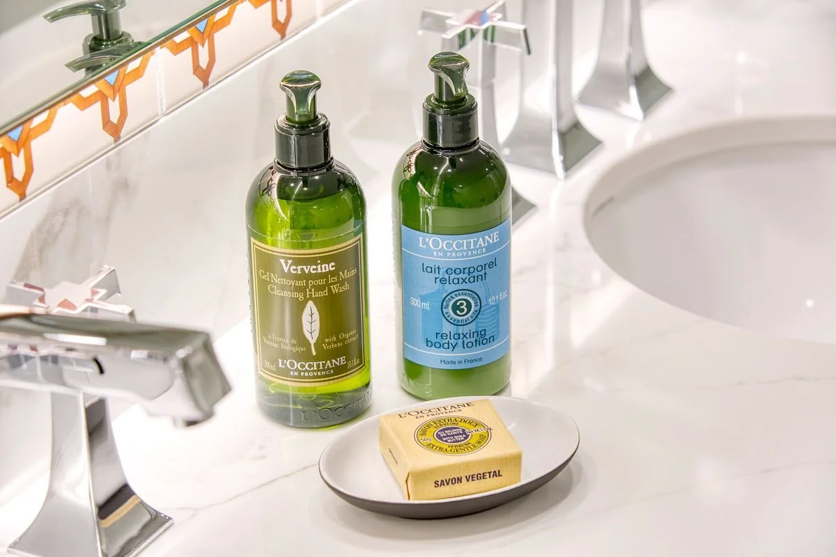 Green toiletry bottles of hand wash and body lotion on a marble bathroom counter, next to a soap bar on a dish, with a mirror and a sink in the background.