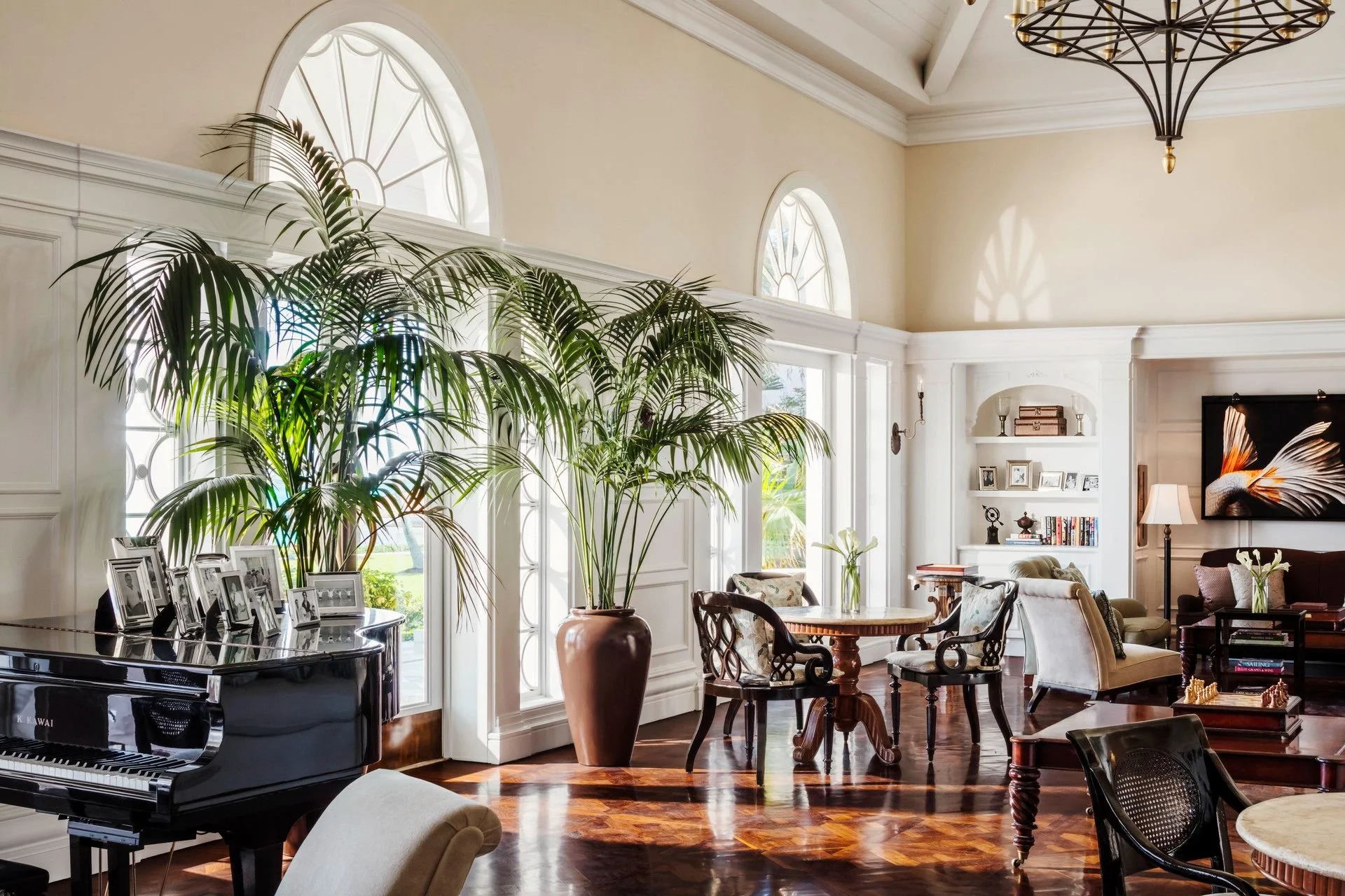 Bright living room with large arched windows, a grand piano with framed photos, tall potted plant, and various seating including chairs and a sofa, decorated with books, lamps, and artwork.