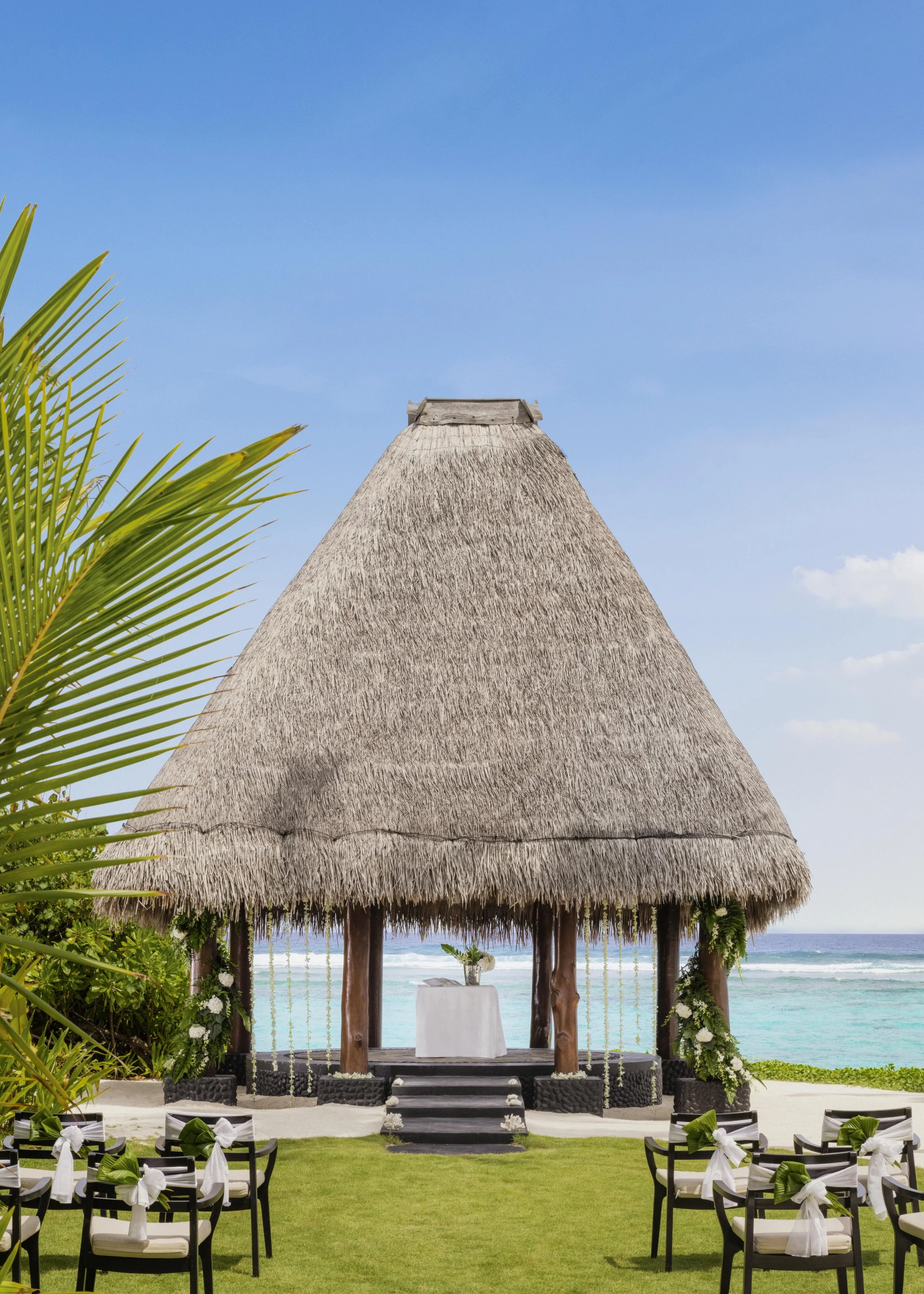Beachside wedding setup with white chairs decorated with green and white ribbons, a thatched roof altar with a white table, tropical plants, and the ocean in the background.