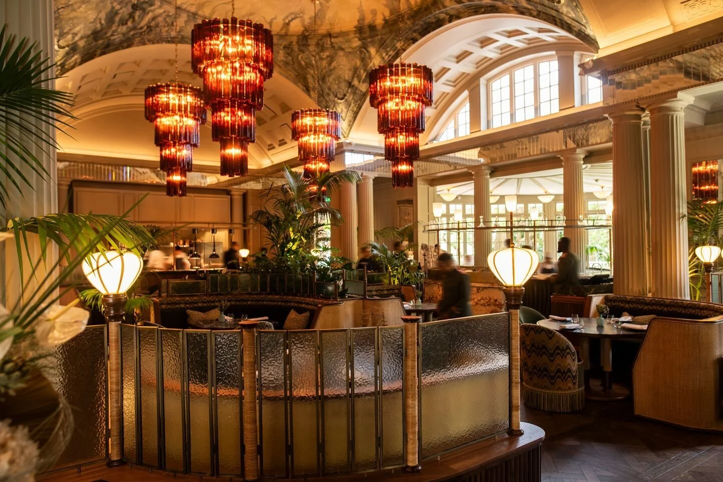 Interior of an upscale restaurant or lounge with ornate ceiling, large windows, red chandeliers, wooden columns, lush green plants, and warmly lit lamps. There are tables with chairs and some people dining or sitting.