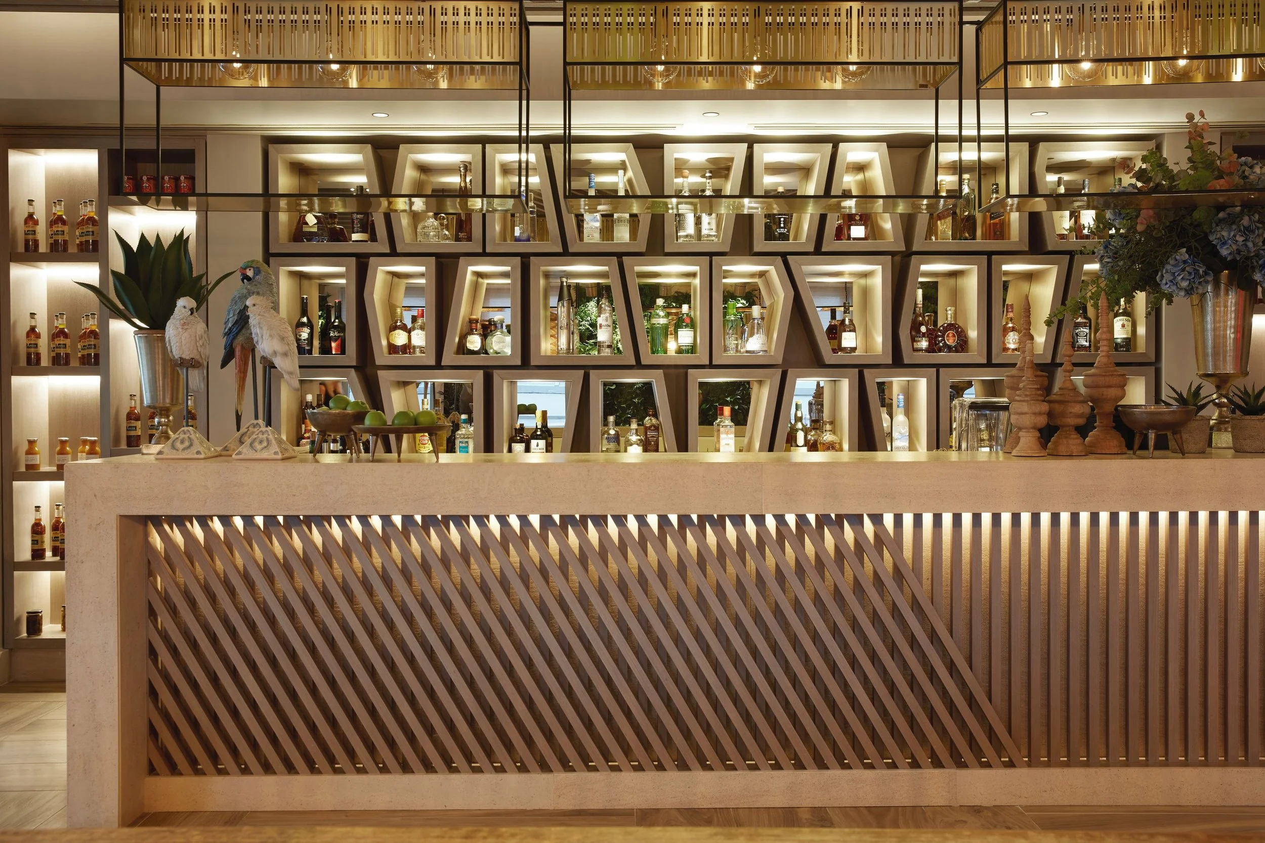 A modern bar setup with illuminated bottles inside geometric wall shelves, a granite counter with decorative pieces including three wooden candlesticks, a potted plant, and colorful artificial birds on a stand.