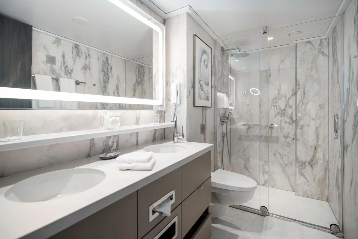 Modern bathroom with marble walls, a double sink vanity, a large illuminated mirror, a glass-enclosed shower area with a rain showerhead, and a framed artwork on the wall.
