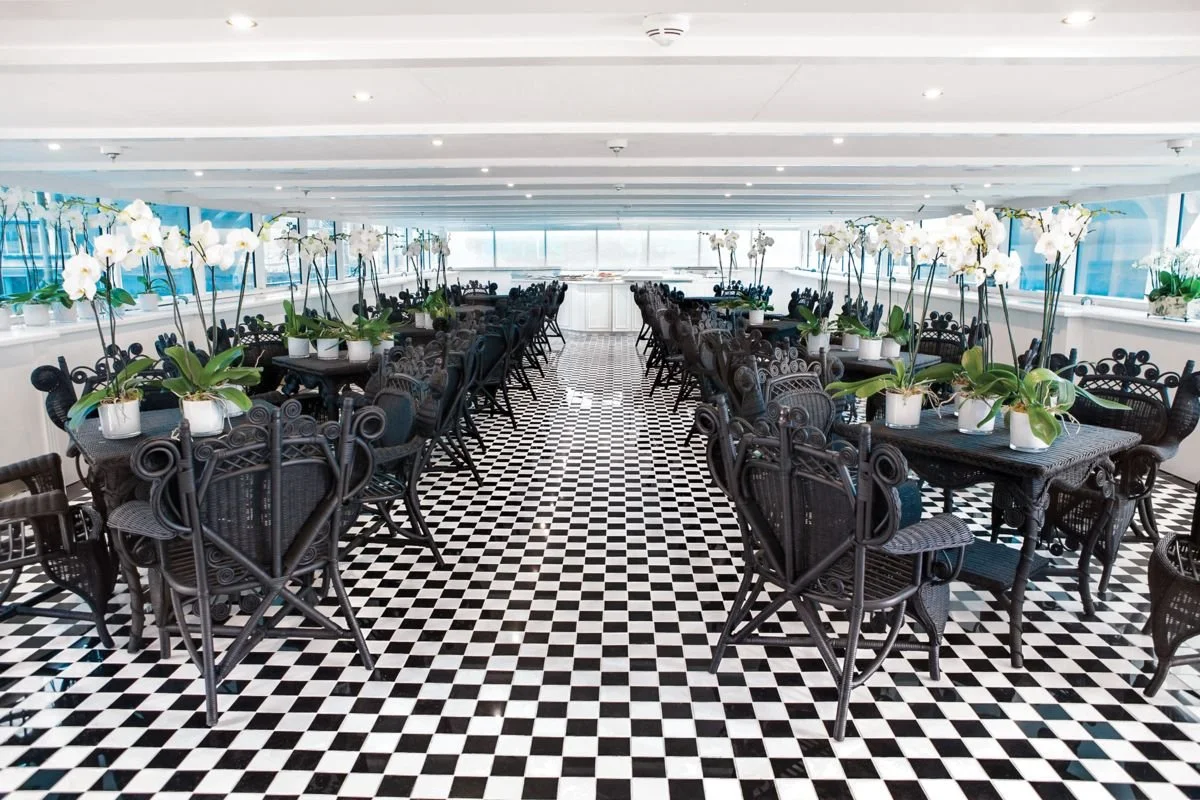 A brightly lit indoor dining area with black wicker chairs and matching tables arranged in rows, decorated with white orchid plants in pots, and a black-and-white checkered floor.