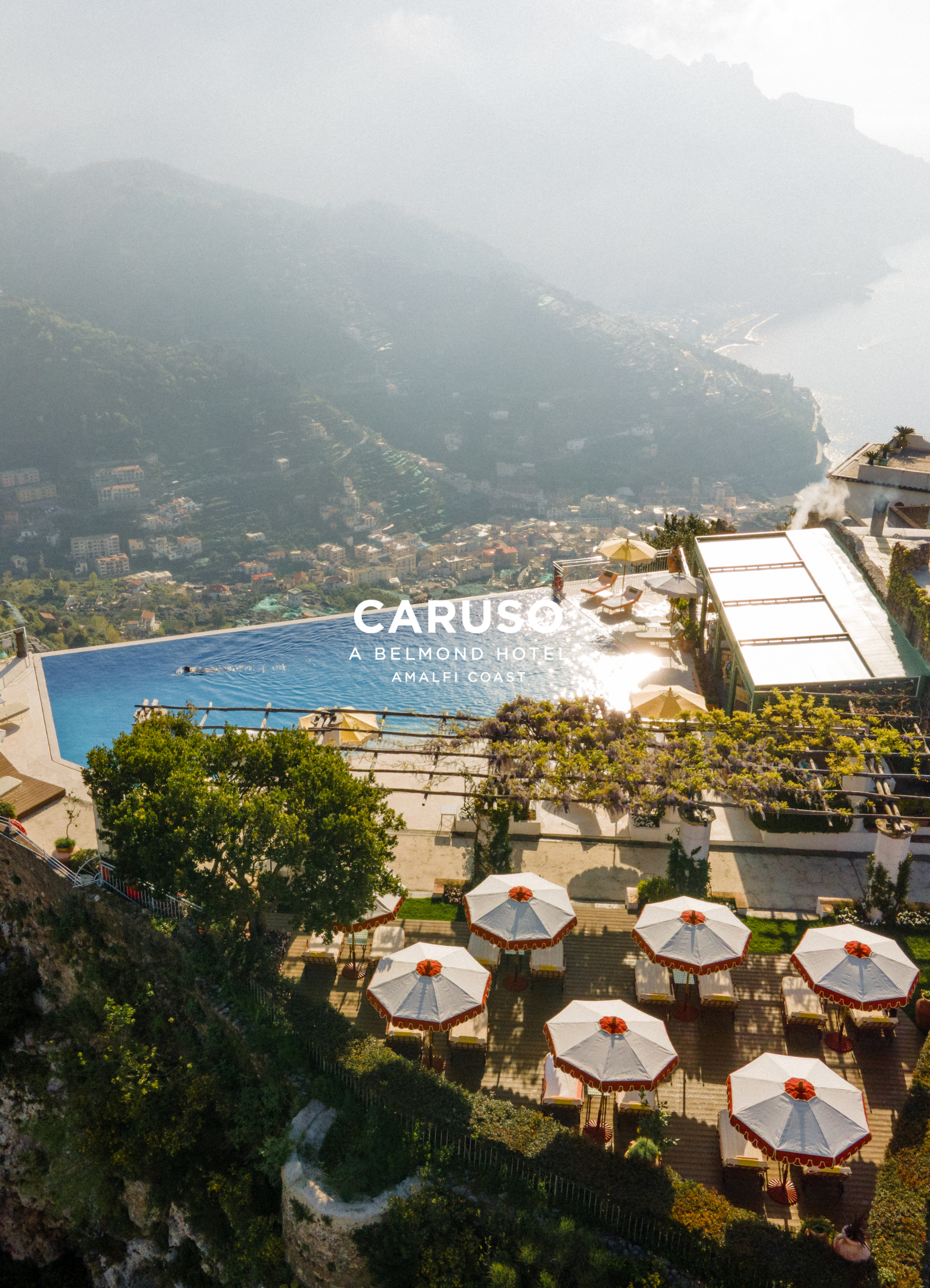 Aerial view of a hotel pool area with white umbrellas, lounge chairs, and a cityscape with mountains and coastline in the background, located in Amalfi Coast.