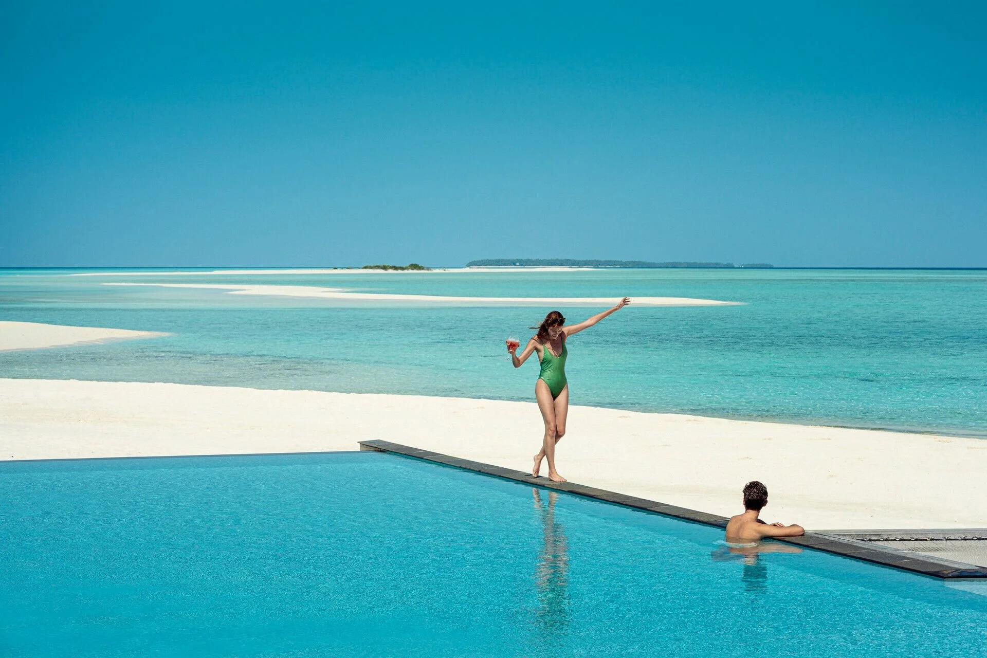 A woman in a green swimsuit balancing on a diving board over a swimming pool, holding a drink, with a woman sitting by the pool and a woman in the water in the background, set against a beach and blue sky.