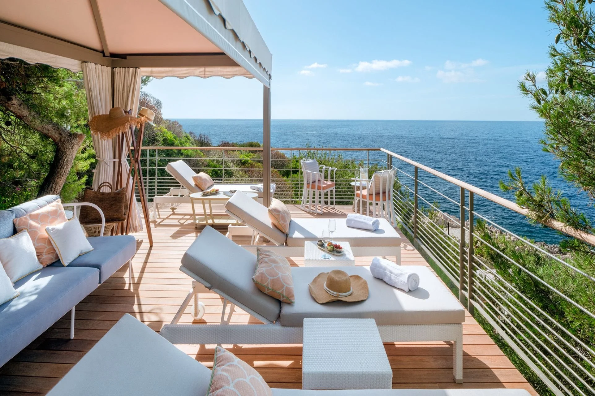 A spacious outdoor patio overlooking the ocean with white lounge chairs, a cushioned sofa, a small table with snacks, and a sun hat, surrounded by trees and greenery under blue skies.