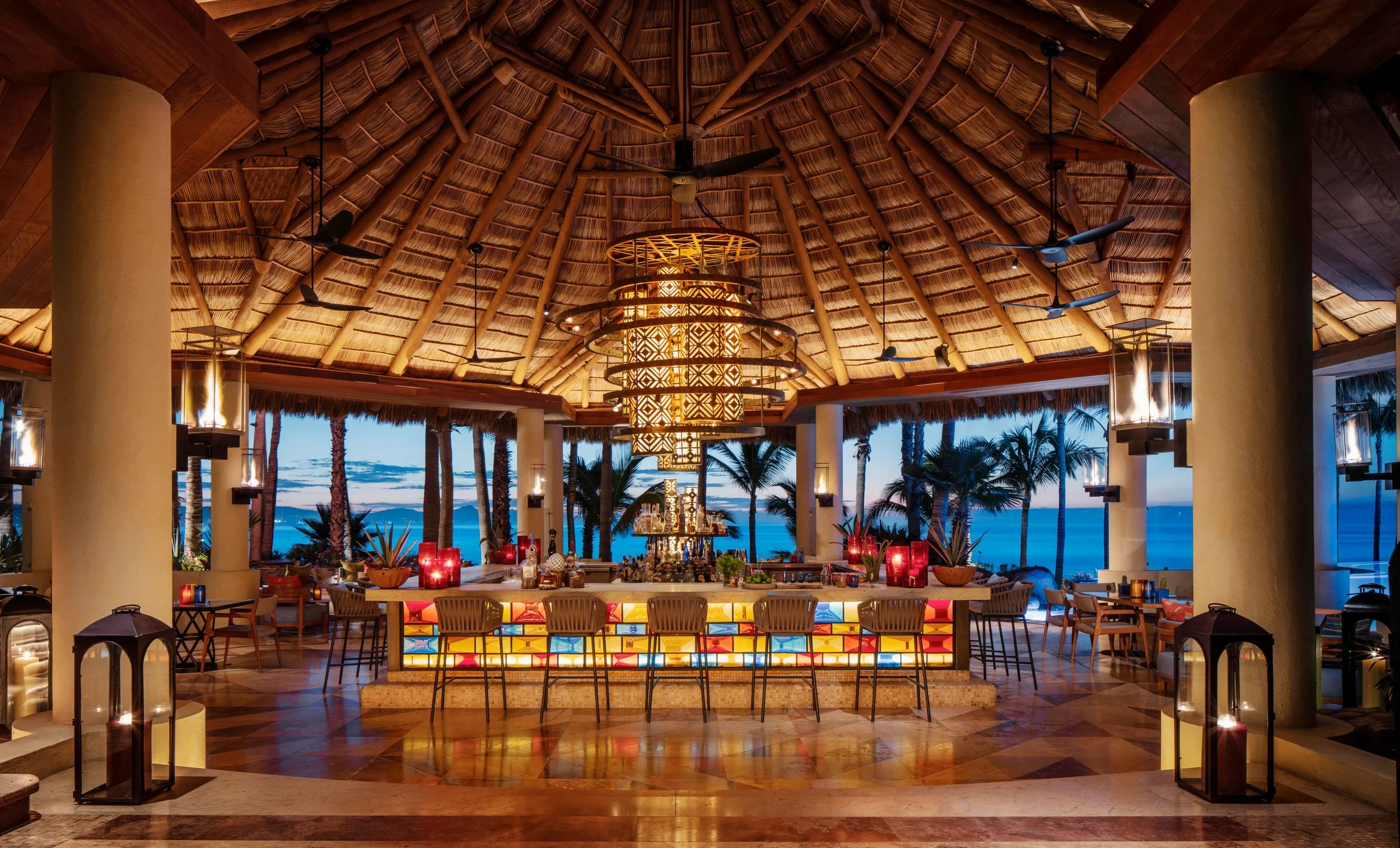 Indoor tropical restaurant with thatched roof ceiling, surrounded by large open windows showing palm trees and ocean view at dusk, decorated with hanging lanterns, a central chandelier, and candlelit tables.