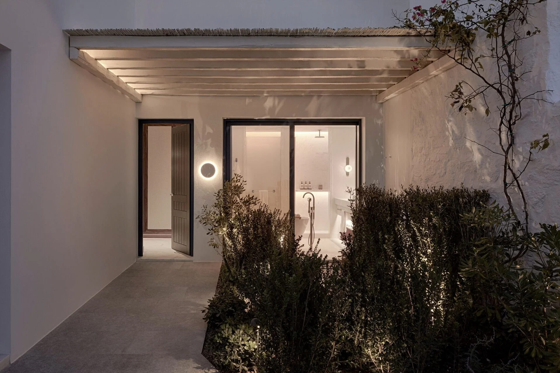 Exterior view of a modern house entrance at dusk with open door, illuminated bathroom interior, greenery, and a vine on the wall.