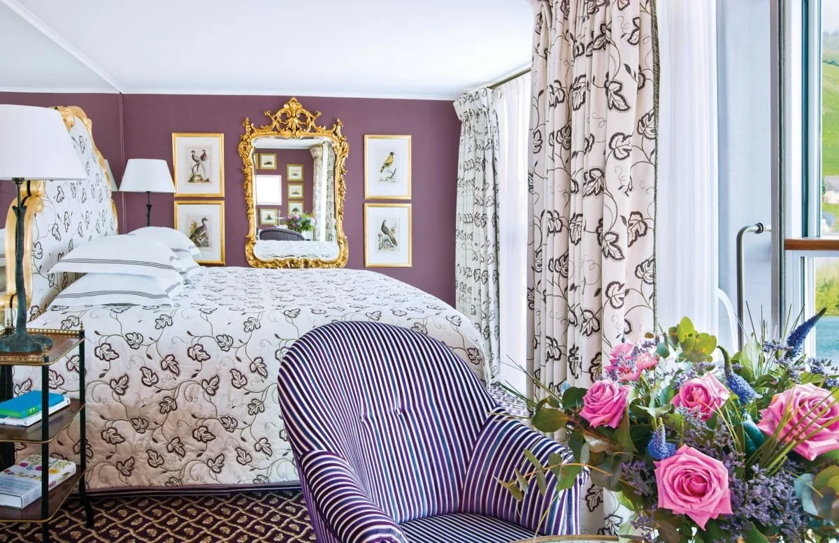 Elegant bedroom with patterned white and black bedding, ornate gold-framed mirror, framed bird artwork on purple wall, striped purple and white armchair, bouquet of pink roses and greenery near large window with floral curtains and a scenic view outside.