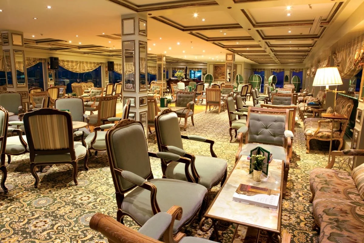 Elegant hotel lounge with numerous chairs and tables, classical decor, ornate carpets, large windows with curtains, and soft lighting.