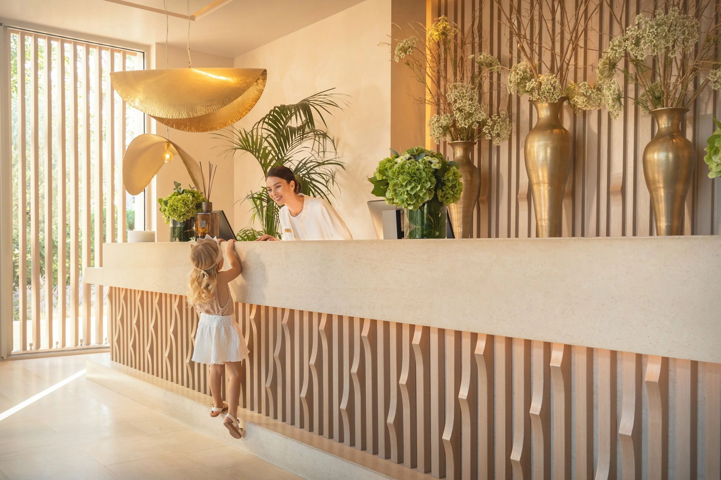 A woman behind a reception desk smiling at a little girl, who is reaching out to touch her. The reception area is decorated with large green plants and tall vases filled with white flowers, with wooden and beige decor accents and large windows letting in natural light.