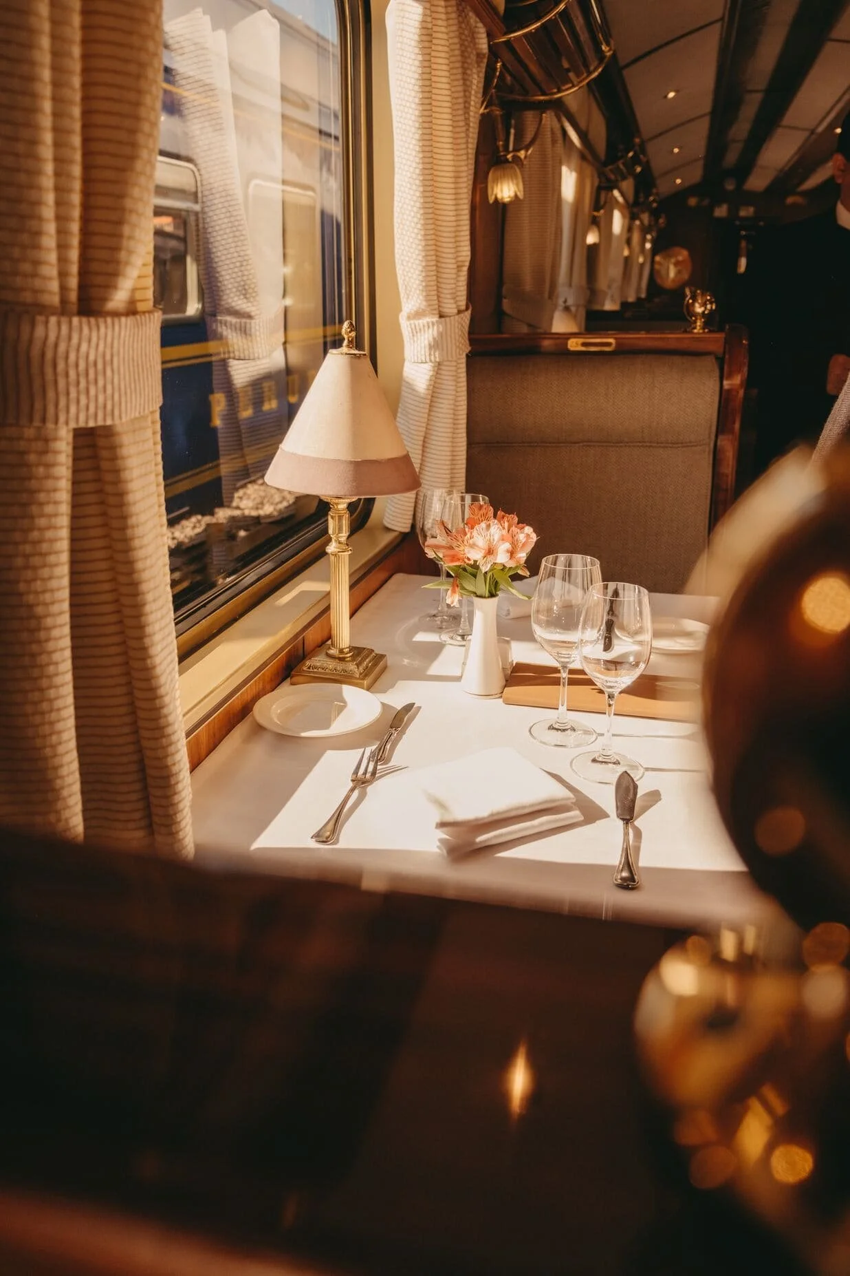 A dining table set with a white tablecloth, a small lamp, a vase with pink flowers, three wine glasses, a plate, silverware, and a folded napkin. The table is near a window with beige striped curtains, and the scene has warm lighting.