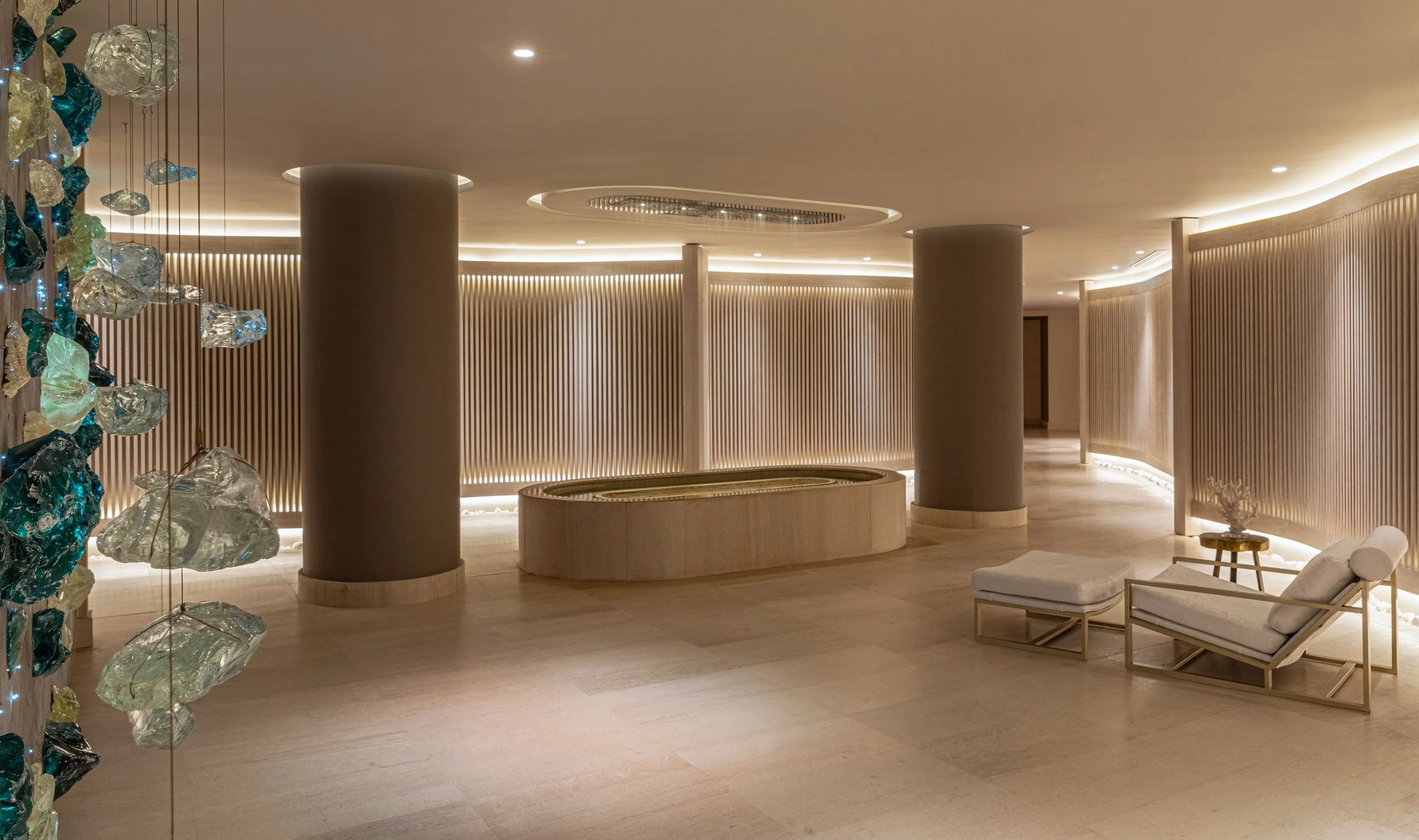 Interior space with neutral tones, featuring decorative lighting, curved wood-paneled walls, a round seating area, and lounge chairs with cushions.