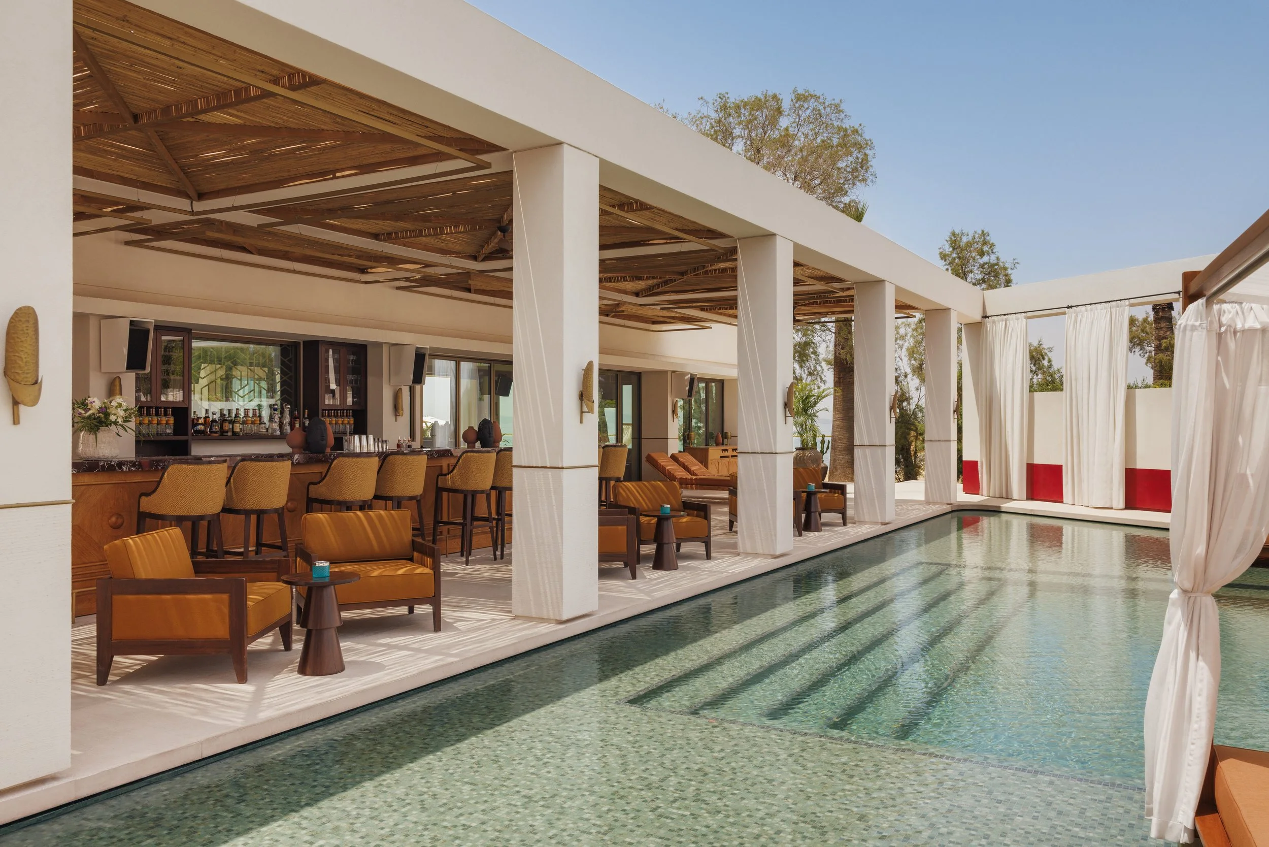Outdoor bar area with seating, pool, and white curtains, under a modern covered structure, with towers and trees in the background.