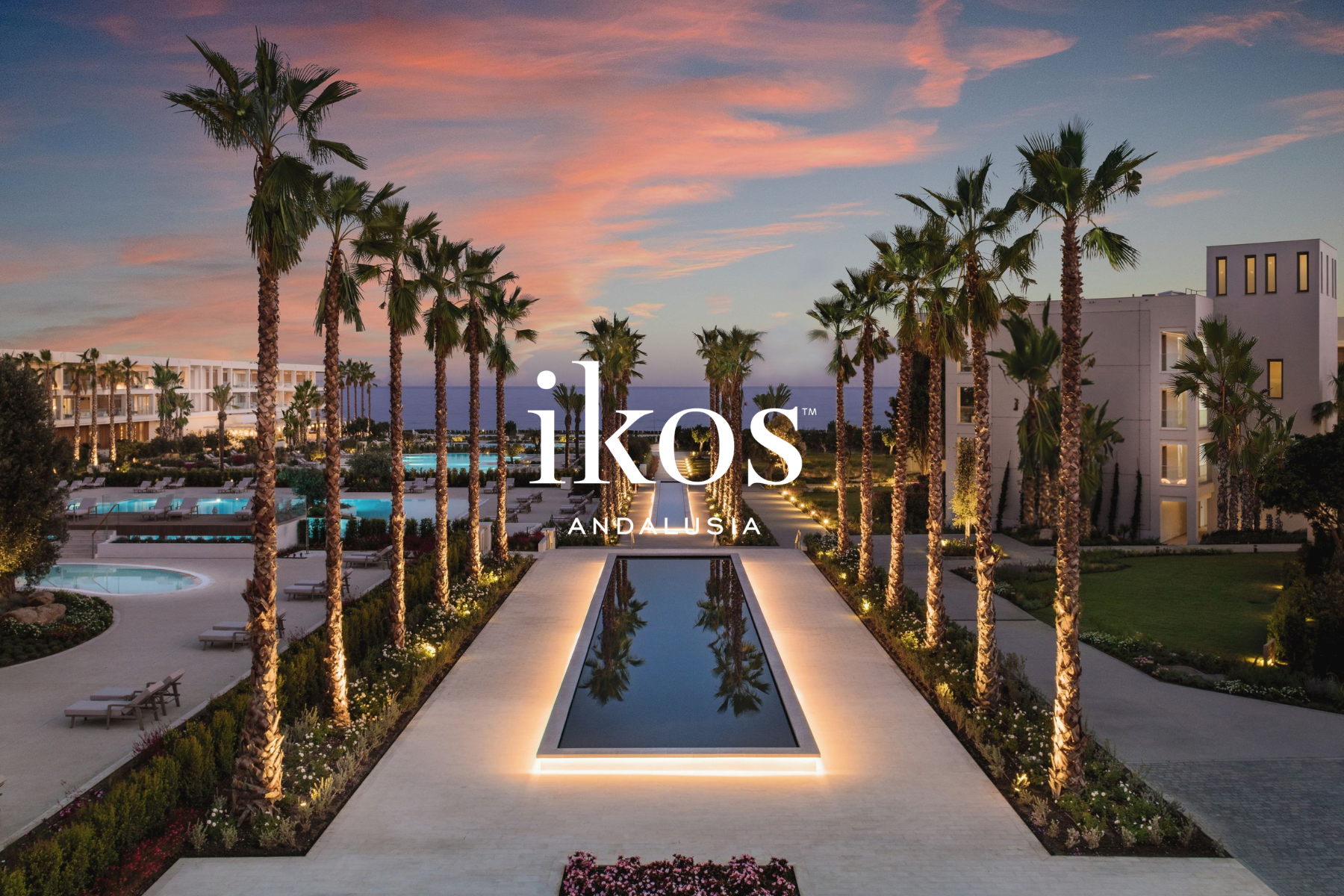 A resort with a long, narrow pool surrounded by palm trees, illuminated walkway, and a garden with flowers, overlooking the ocean at sunset with pink and blue sky.