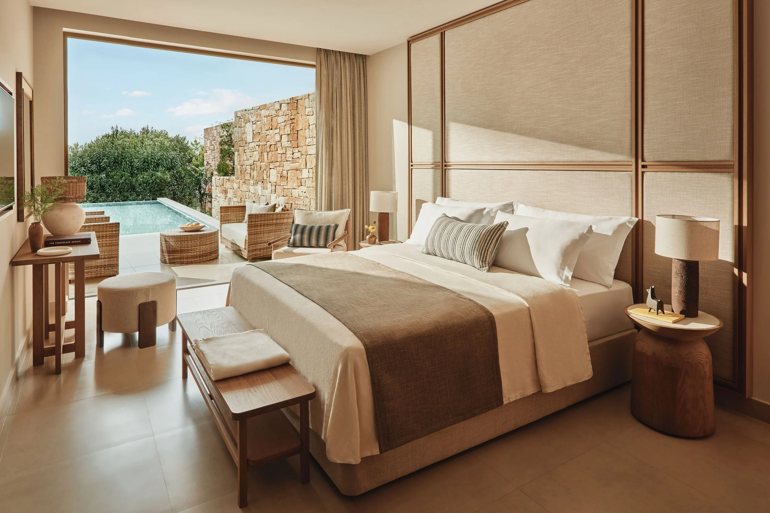 A hotel room with a large bed, nightstand, and a view of an outdoor pool and lush greenery through a sliding glass door with beige curtains.
