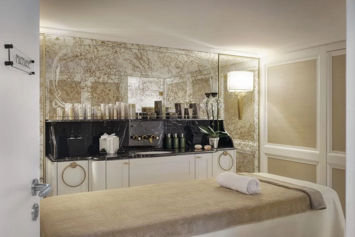 A luxurious massage room with a massage table covered with a beige blanket and a rolled towel on top. The room has a black marble counter with candles, a tissue box, and decorative items. There is a large mirror with a gold frame on the wall, a wall-mounted light fixture, and ornate wall paneling in the background.