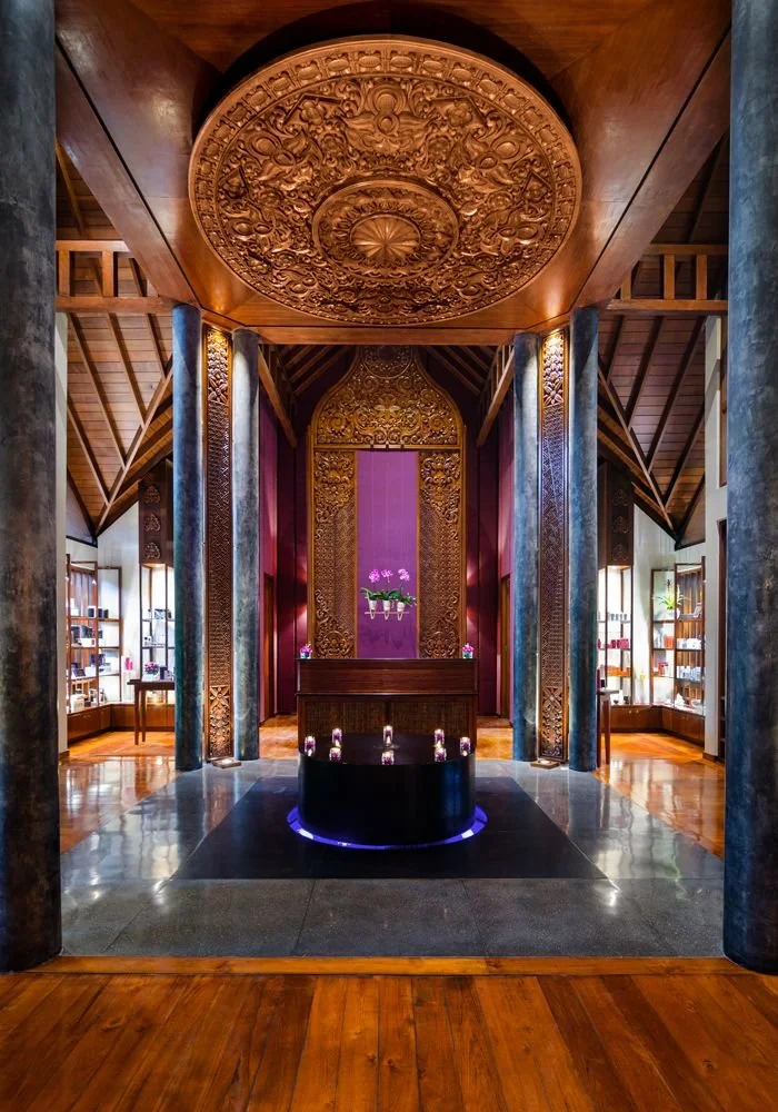 Interior of a room with intricate wooden carvings on the ceiling and walls, showcasing Asian-inspired design. There are four columns with decorative details, and purple orchids on a small platform in the center. Windows with bookshelves and natural light are visible on either side.