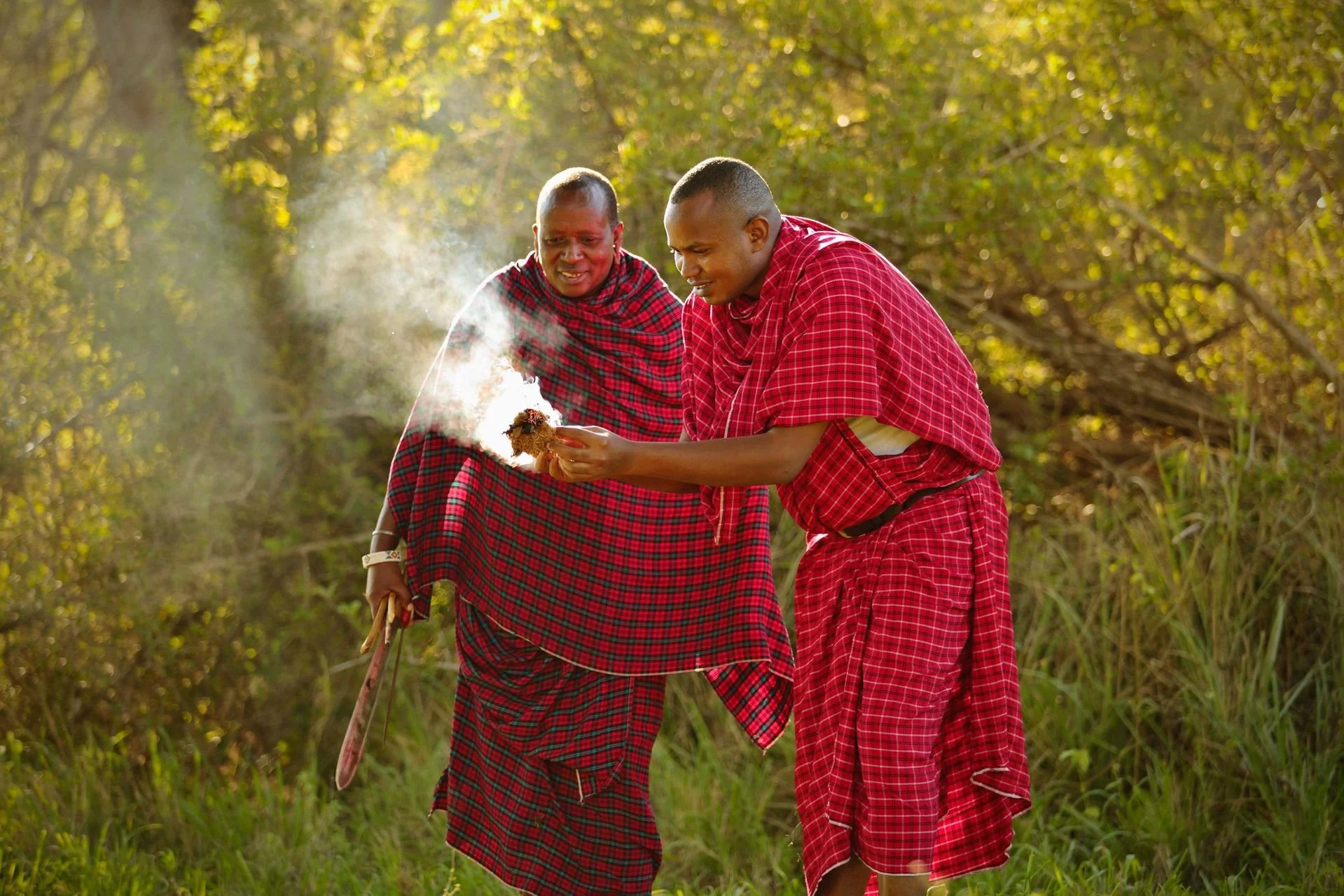 Two Maasai men wearing traditional red and black shúkà Cloak inspecting a piece of termite mound in a sunlit outdoor setting with green trees in the background.