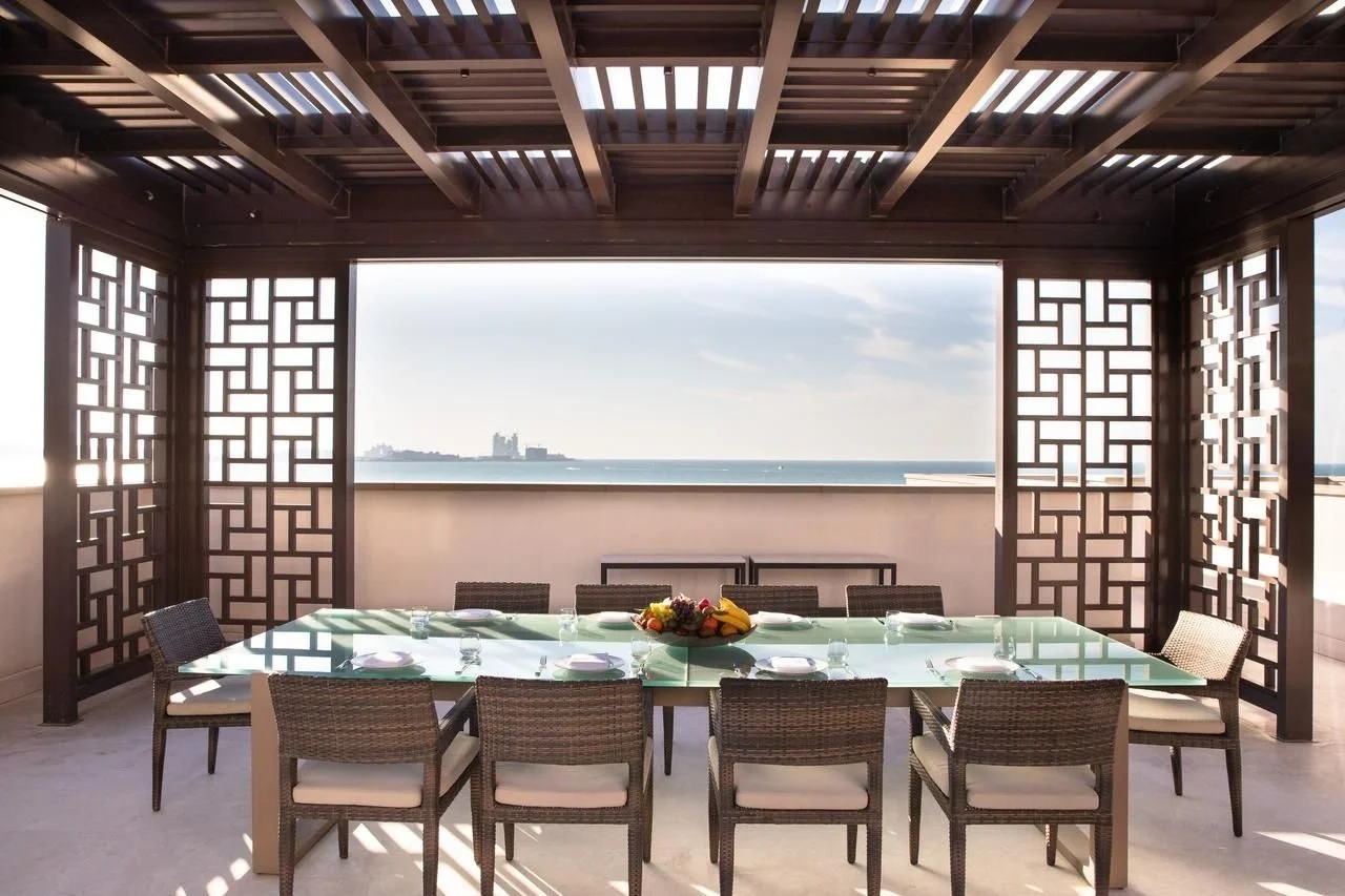 Outdoor dining area with a glass table and wicker chairs, overlooking the ocean, with a wooden pergola providing shade.
