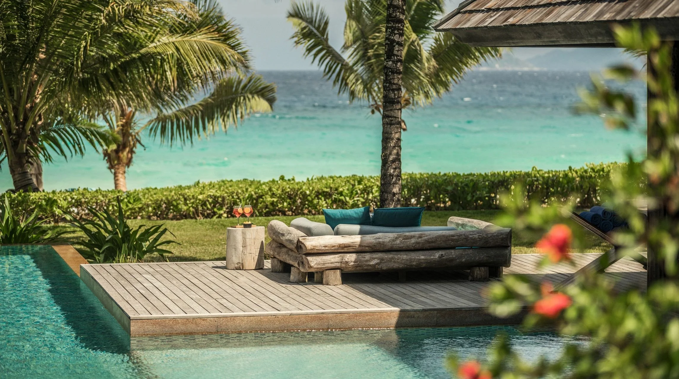 A poolside seating area with a view of the ocean, tropical trees, and lush greenery in the background. The seating includes a rustic wooden bench with turquoise cushions, a small stone table with two drinks, and rolled towels.