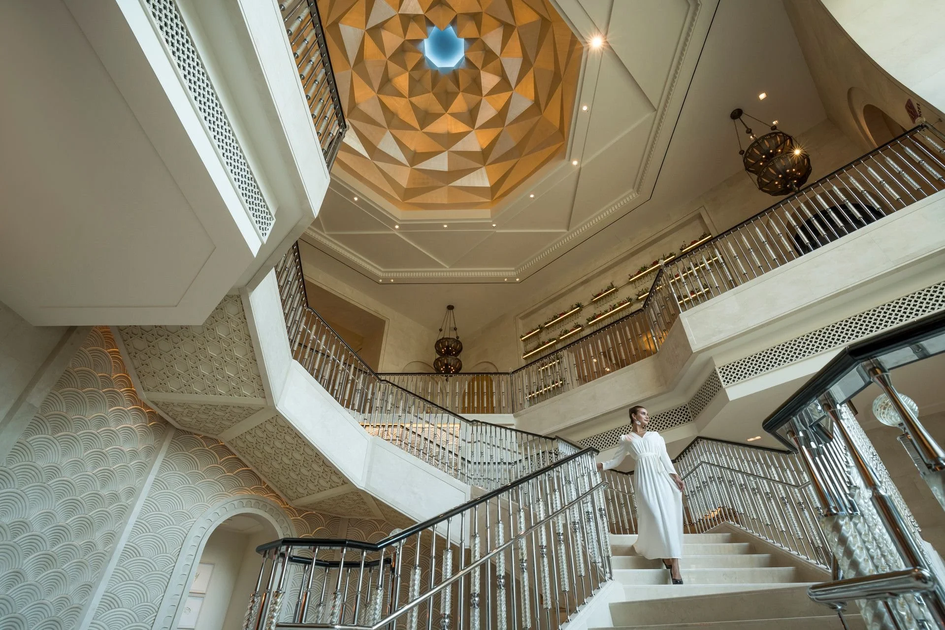 Interior view of a grand hotel or building with multiple floors, featuring intricate woodwork on the ceiling, elegant lighting fixtures, and a woman in a white dress standing on a staircase railing.