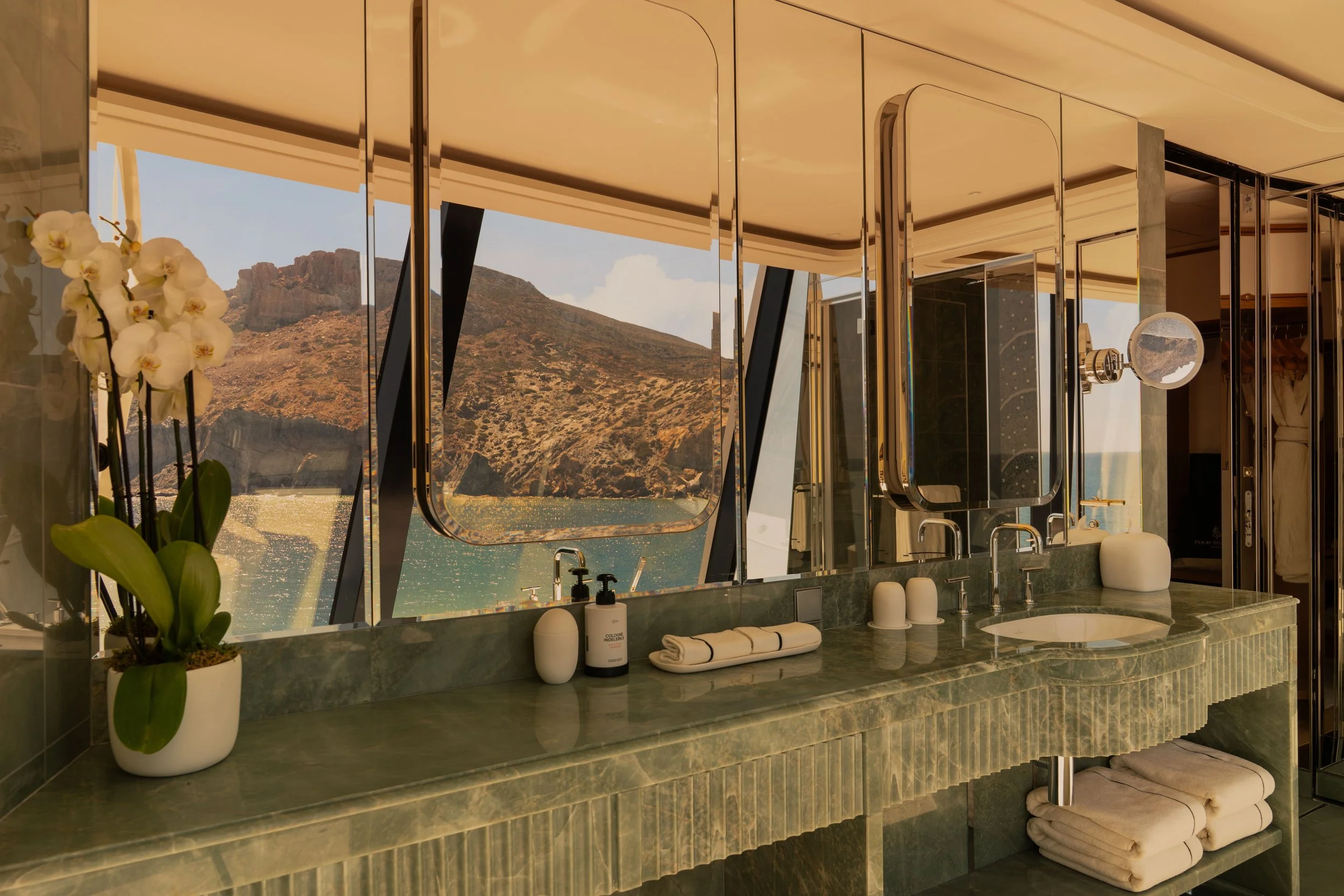 A luxurious bathroom with a large mirror and a view of a mountain landscape through window panels. There is a green marble countertop with a sink, white towels, and a potted orchid plant.