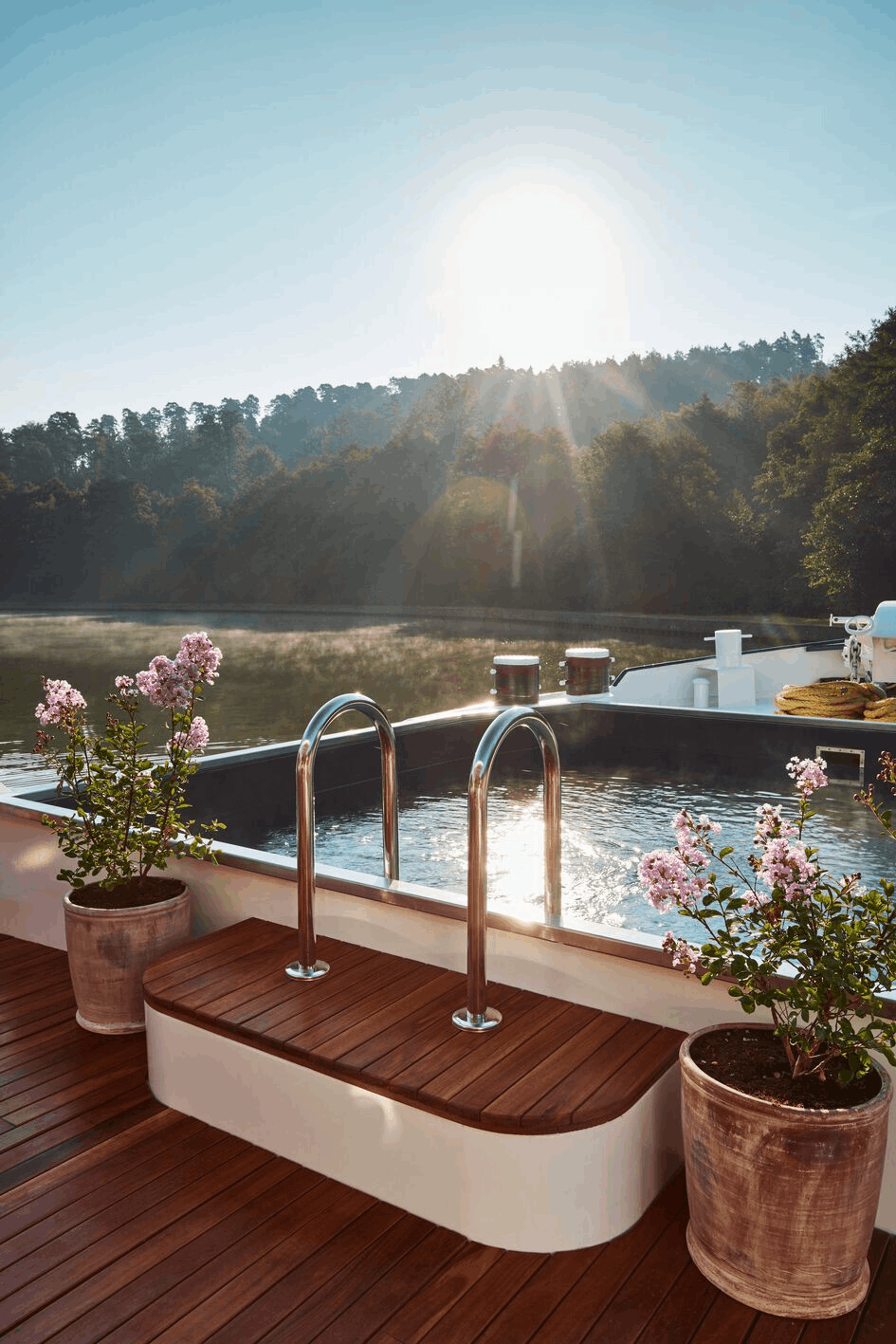 A peaceful lakeside scene at sunset with a hot tub on a wooden deck, potted pink flowering plants, and a mountain landscape in the background.