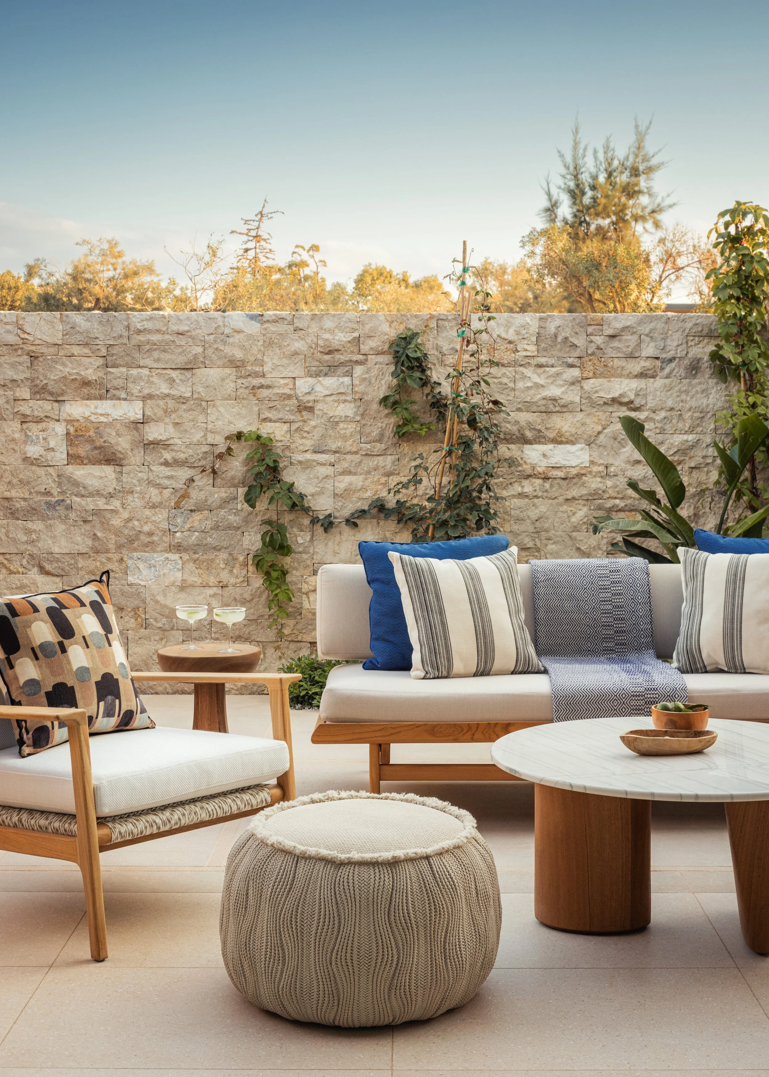 Outdoor patio with beige stone wall, various seating including a white sofa with blue and striped pillows, wooden chairs, a round marble-top table, and a knitted pouf. Green plants and trees in the background, clear sky, warm and inviting setting.
