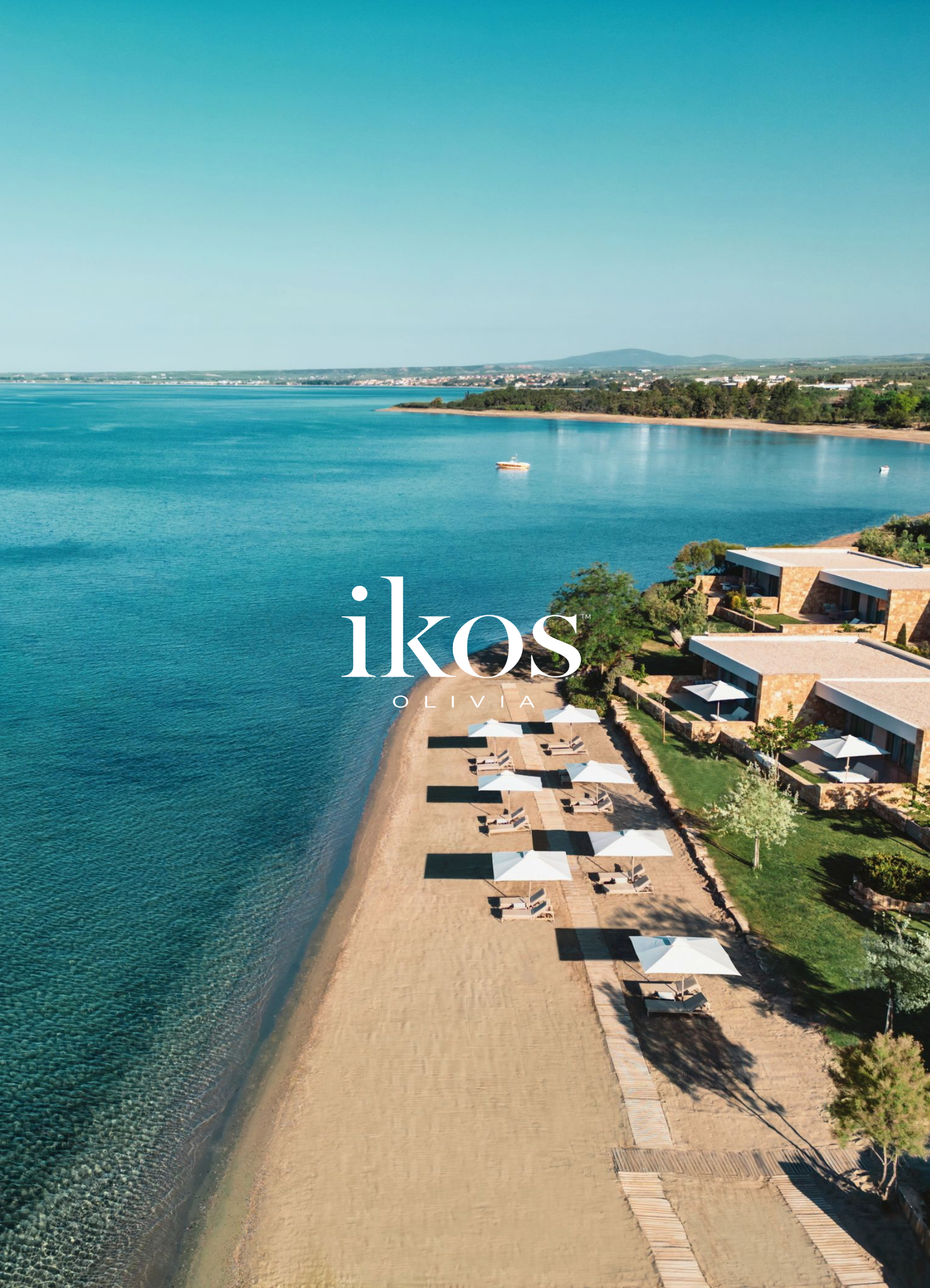 Aerial view of a beach with a shoreline, lounge chairs under white umbrellas, modern villas, and calm blue water with boats, overlooking distant hills and a clear sky.