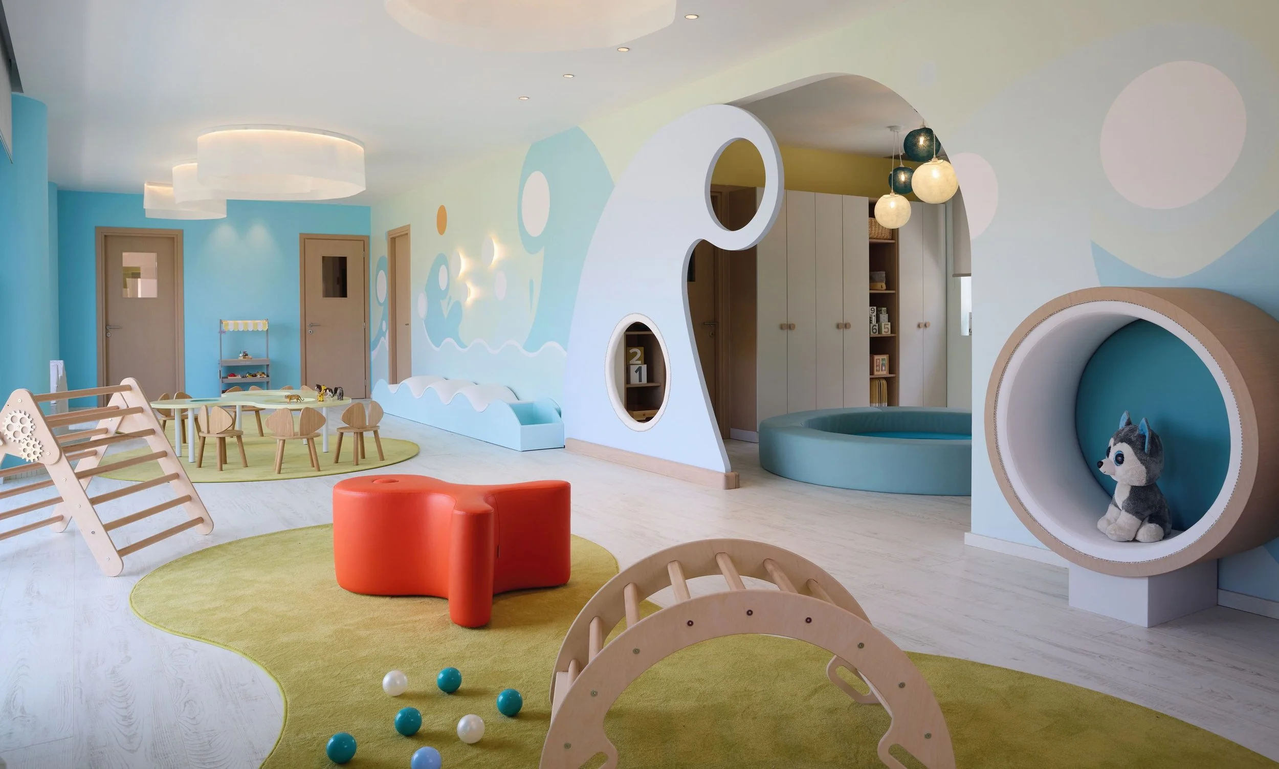 Colorful children's playroom with soft foam furniture, play golf, tables, chairs, and a cozy nook with a plush toy husky. Bright yellow, blue, and green accents with natural wood furniture and modern lighting.