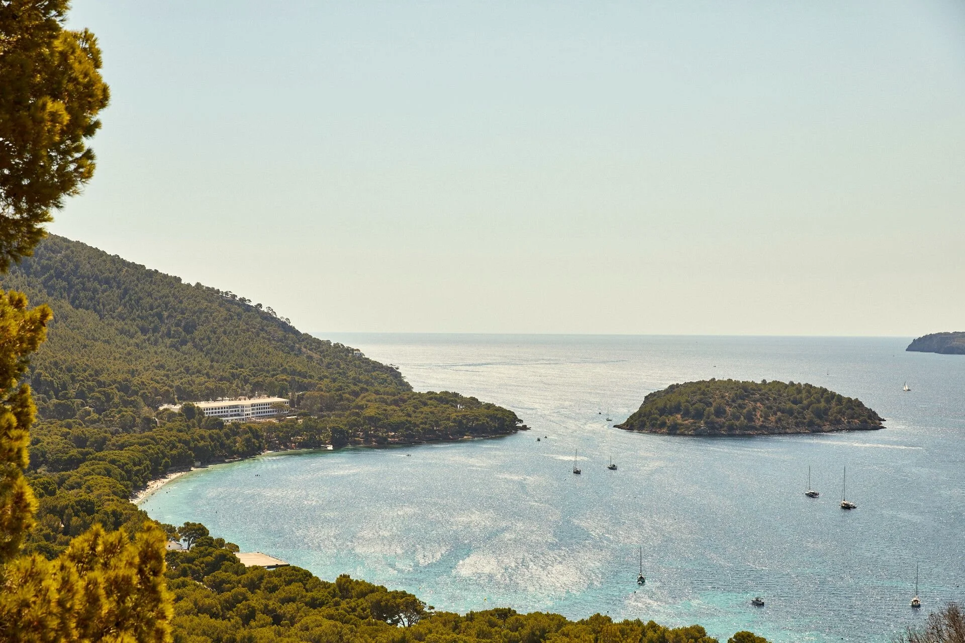 A scenic view of a bay with sailboats, surrounded by forested hills, with a distant horizon and a boat sailing.