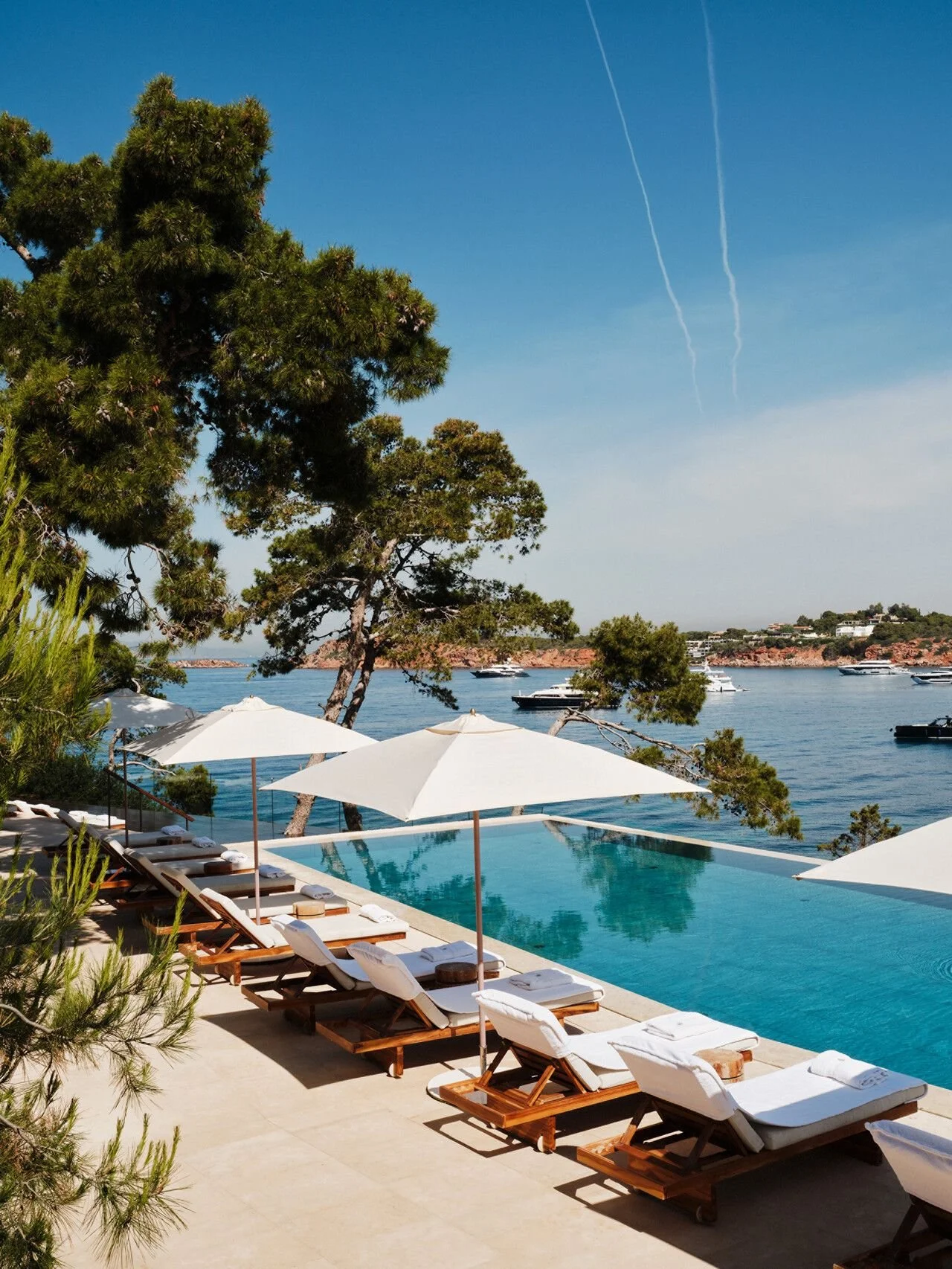 Poolside area with white umbrellas and lounge chairs overlooking a calm ocean with boats, trees, and hills in the background under a clear blue sky with contrails.