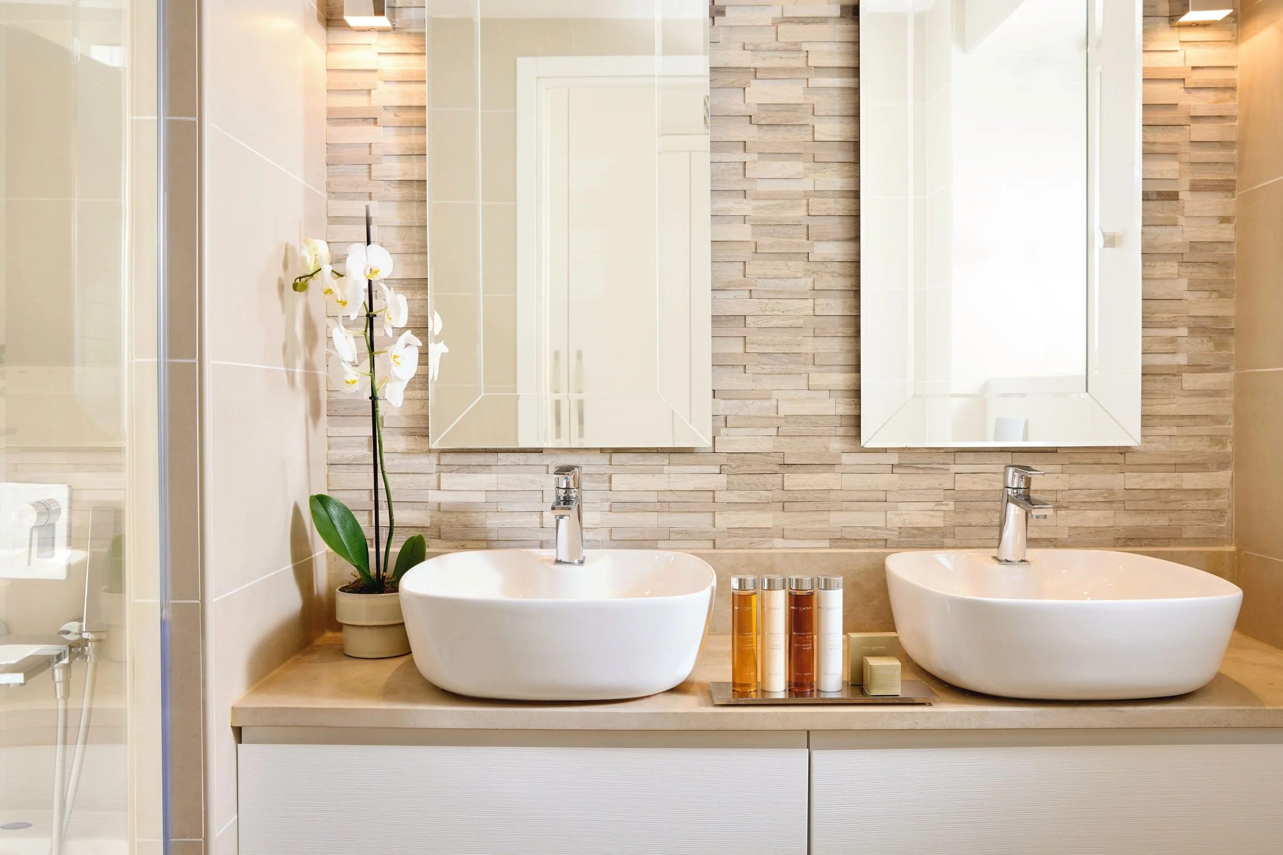 Double vanity bathroom with two white vessel sinks, a large mirror, a potted orchid, and toiletries, with wood tile accents.