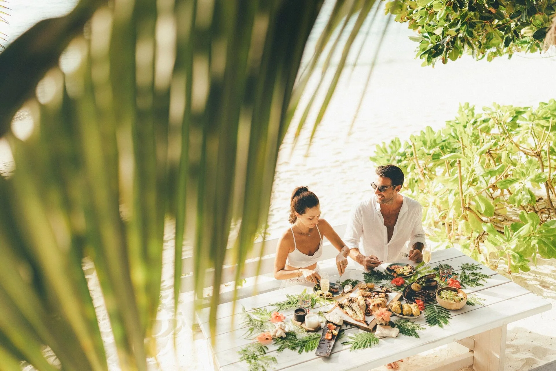 A group of people enjoying a tropical outdoor meal under lush green trees, with a variety of food on a large white table.
