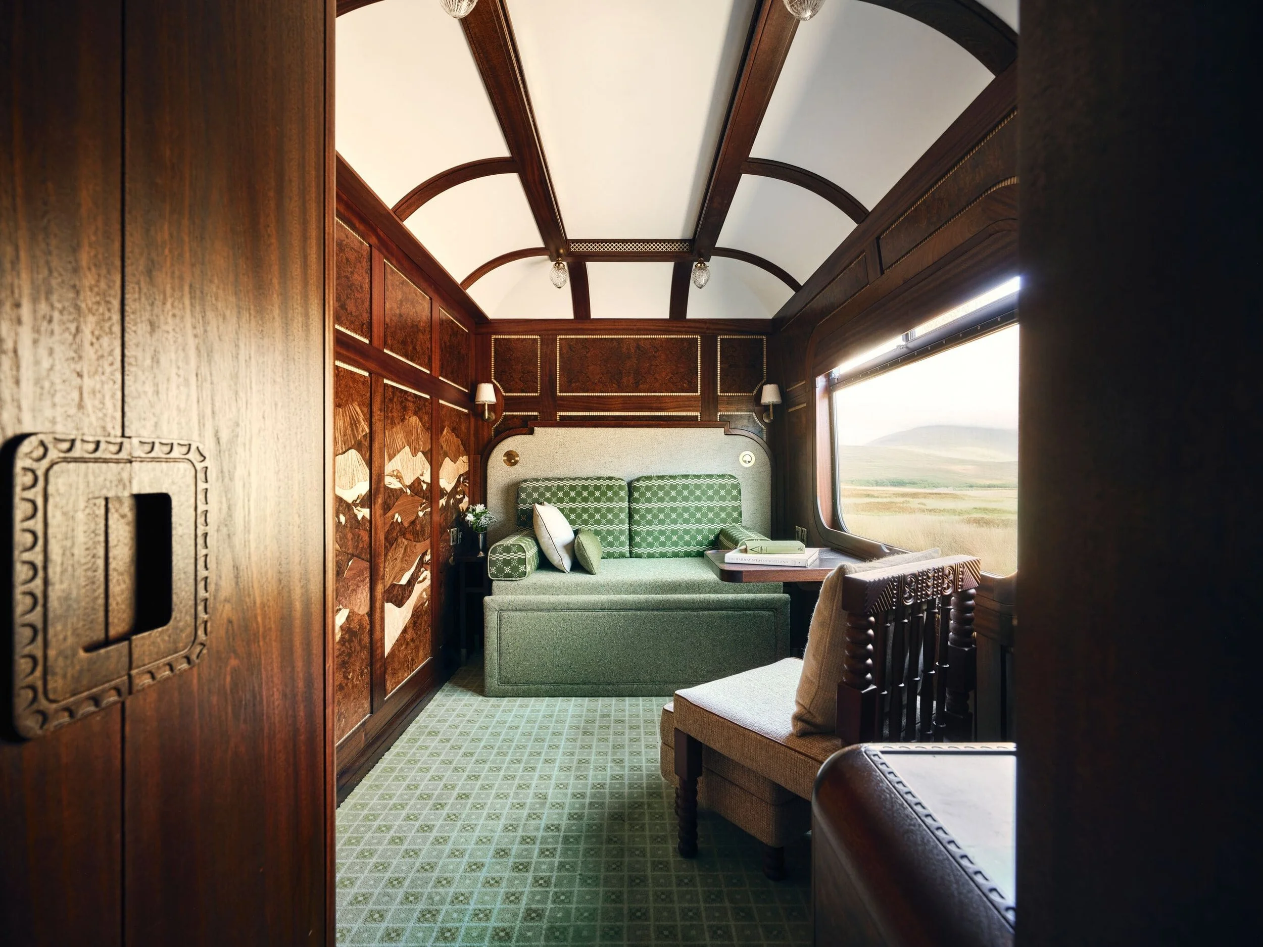 Interior of a vintage train car with wood-paneled walls, a green patterned sofa, and a view of open fields through large window.