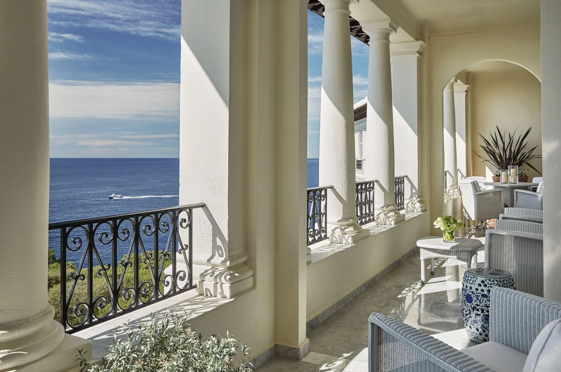 View of a balcony with white columns and ornate black iron railing overlooking the ocean, with a boat sailing in the water, and outdoor seating with white wicker furniture and decorative plants.