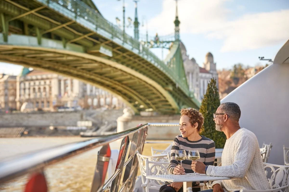 A couple drinking wine on a boat by a river, with a green bridge overhead and city buildings in the background during daytime.