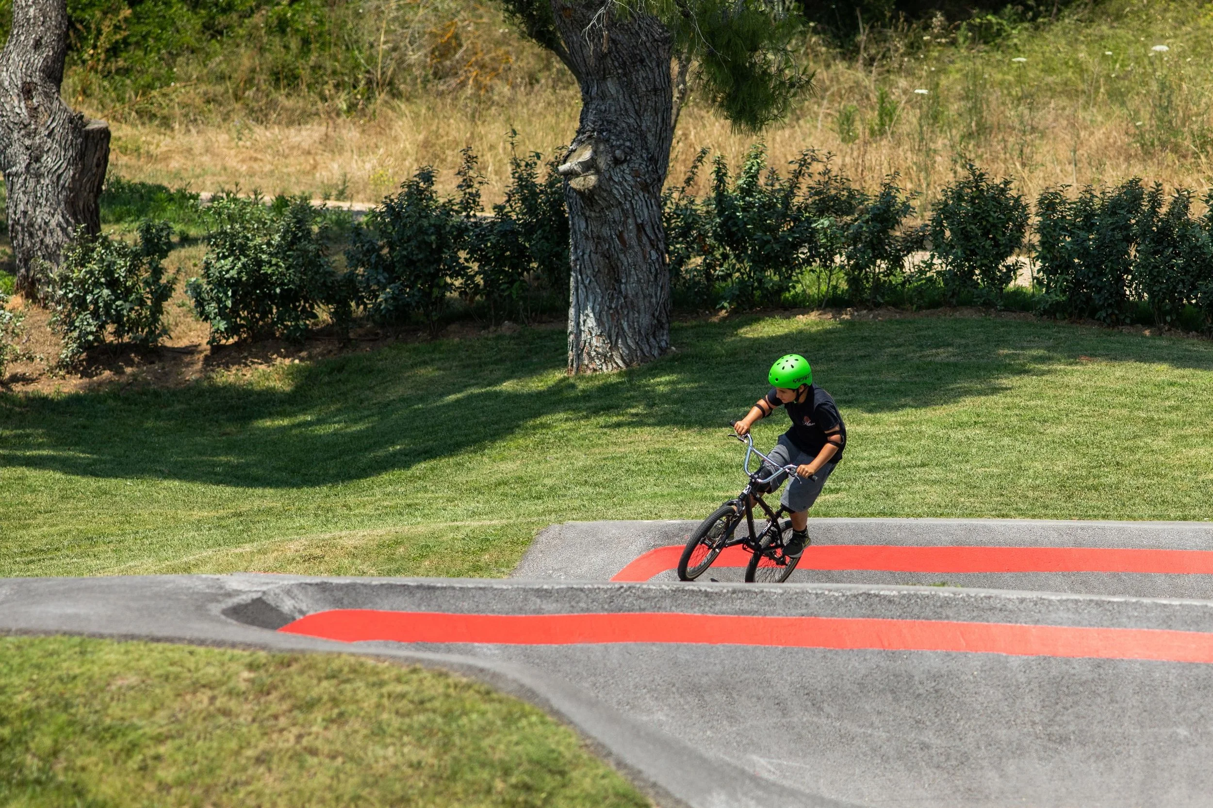 A child wearing a green helmet riding a bicycle on a paved track with a red section, surrounded by grass and trees.