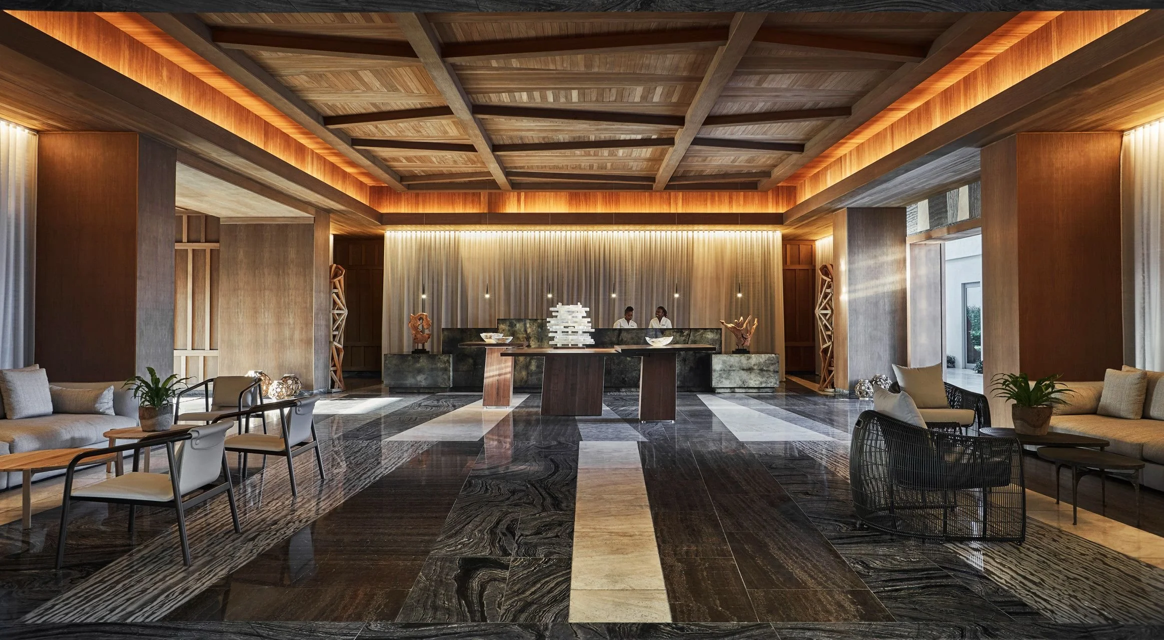 Luxurious hotel lobby with wooden ceiling, modern reception desk with two staff members, and various seating areas with sofas and chairs, decorative sculptures, indoor plants, and marble flooring.