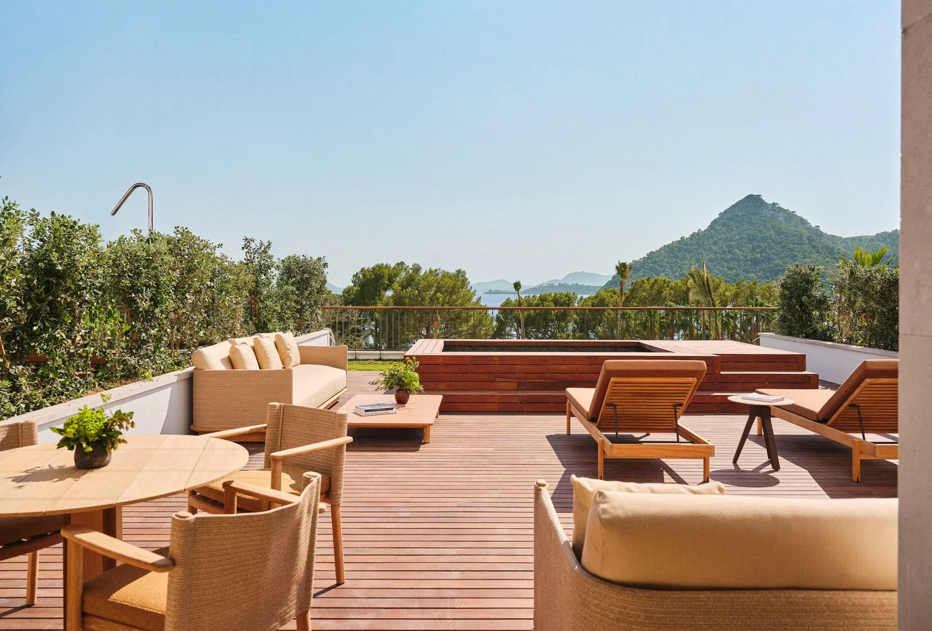 A sunny outdoor patio with wooden decking, comfortable outdoor furniture including chairs and a sofa, potted plants, and scenic views of green trees and distant mountains under a clear blue sky.