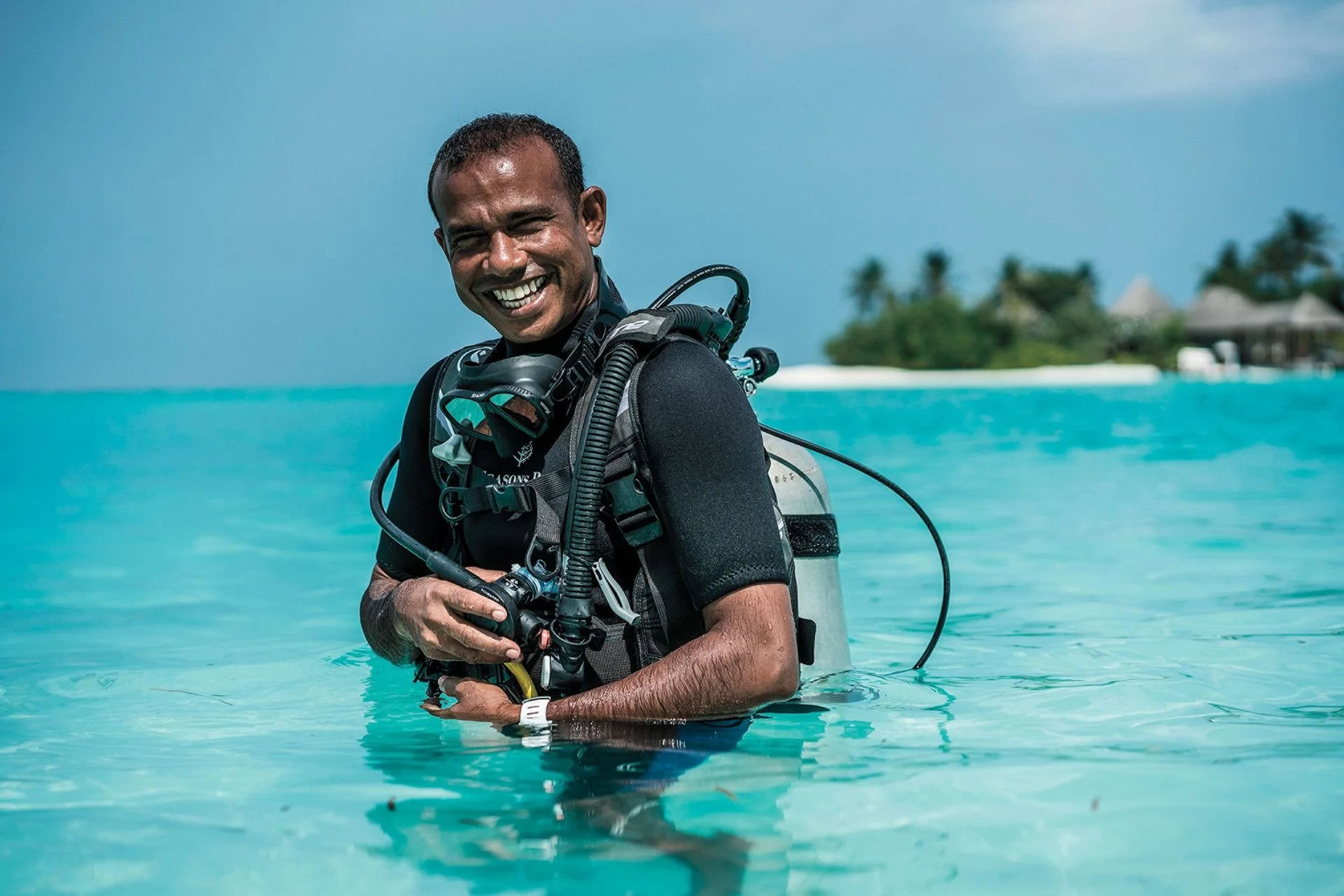 A man in scuba gear standing waist-deep in clear turquoise ocean water, smiling, with a tropical island with huts and palm trees in the background under a blue sky.